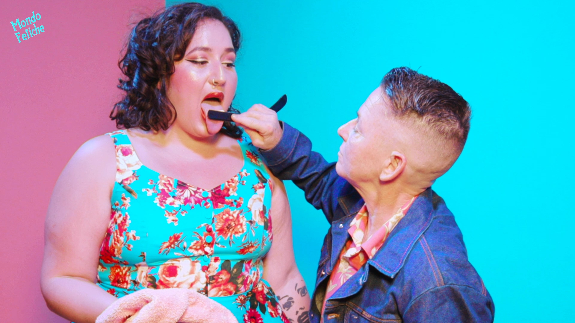 Image shows a pin-up styled person with her tongue out, while another person wearing a denim jacket puts their shaving razor on the others tongue making her lick it clean.