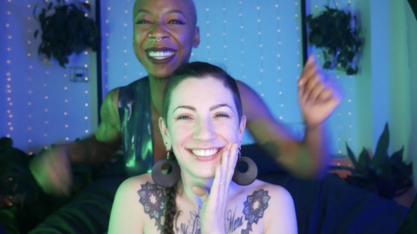 Porn Performer Ashley Paige, who has a shaved head and wears a leather vest, stands behind porn performer Corey More, who also has a shaved head and wears large, circular earrings. They both smile.