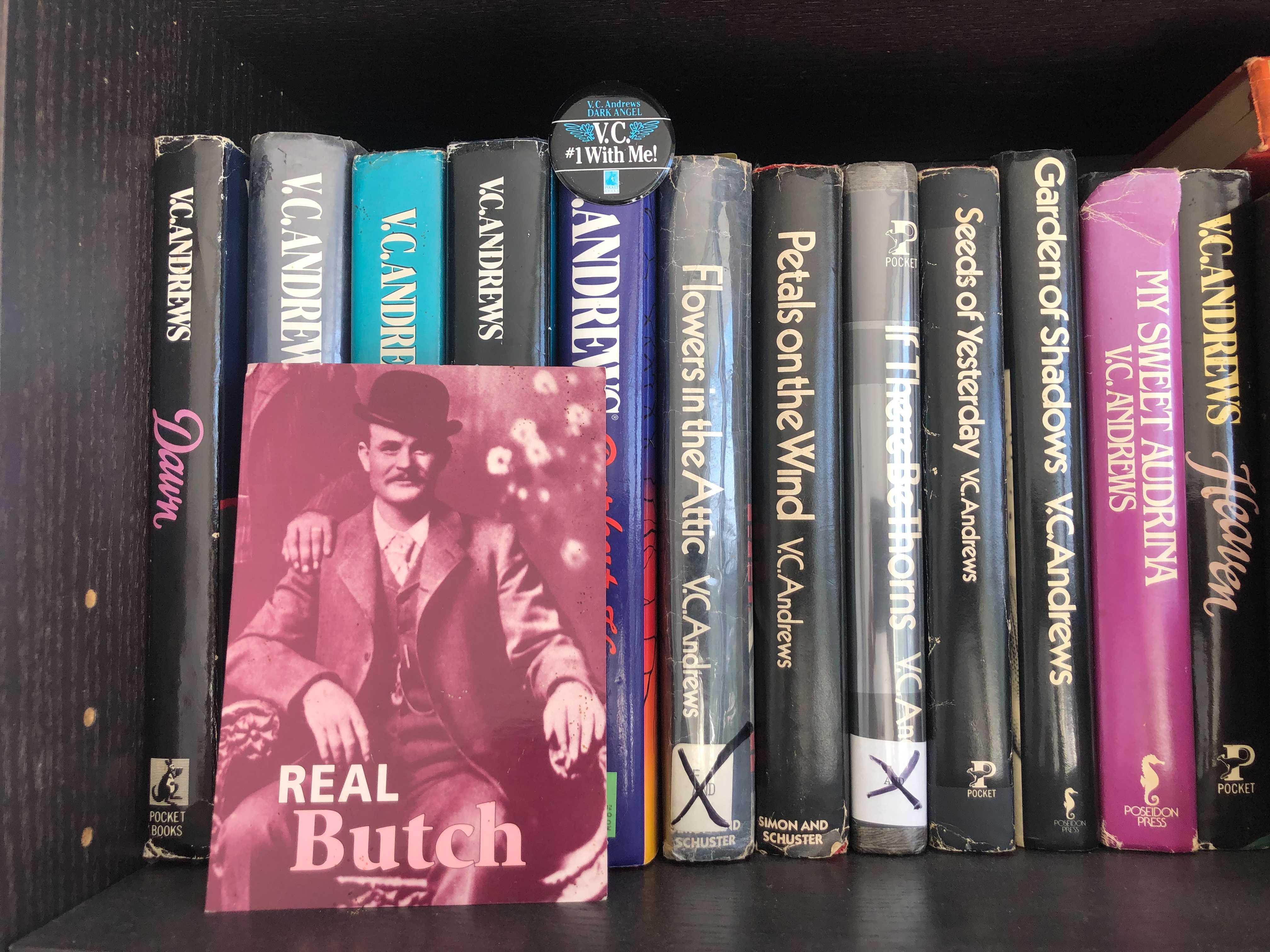 a shelf of V.C. Andrews books with a "real butch" postcard