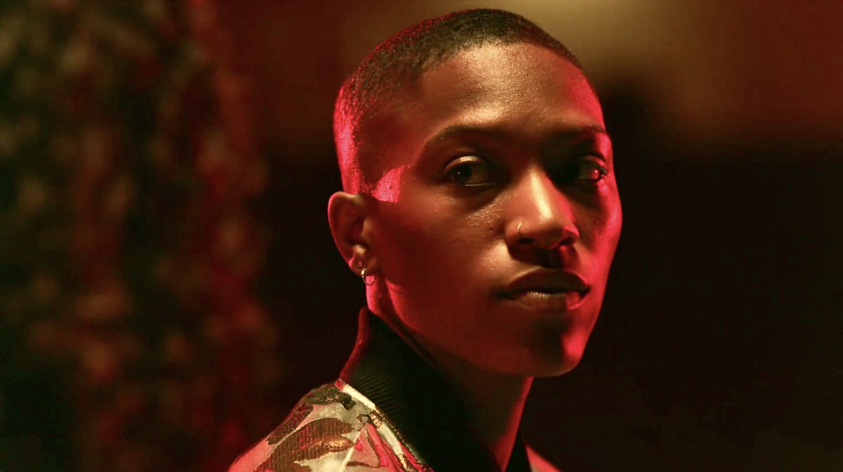 Image shows Hattie, a Black masc lesbian, looking very seriously over her shoulder. Hattie is lit in red undertones.