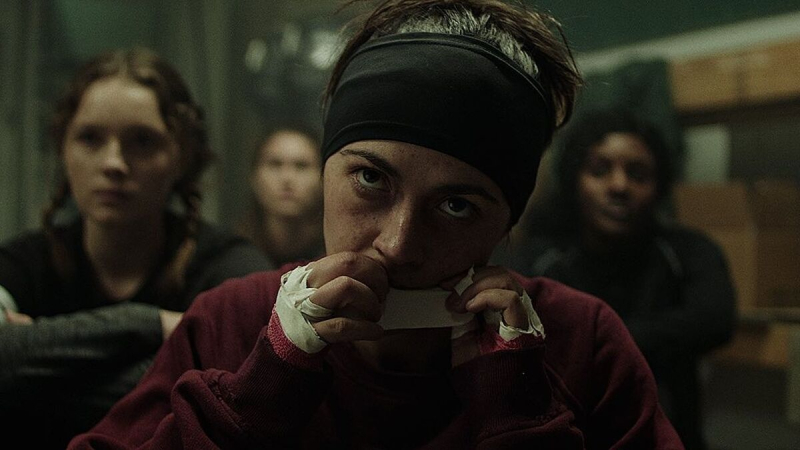 lesbian movies 2021 list still: Isabelle Fuhrman tears athletic tape with her teeth as she looks up at her coach while sitting with her teammates. She wears a black headband and red sweatshirt, her bloodied hands already wrapped in tape.