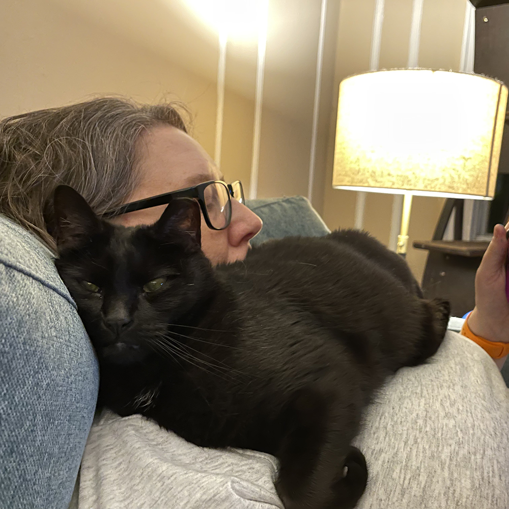 Beth March, a tiny all-black cat, lays on top of Heather's chest, tucked under her chin, as she plays her Nintendo switch on the couch.