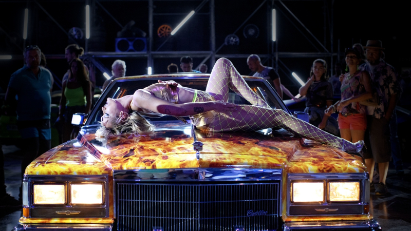 Agathe Roussel in a gold bra and neon yellow fishnets leans back on the hood of a fire painted car.