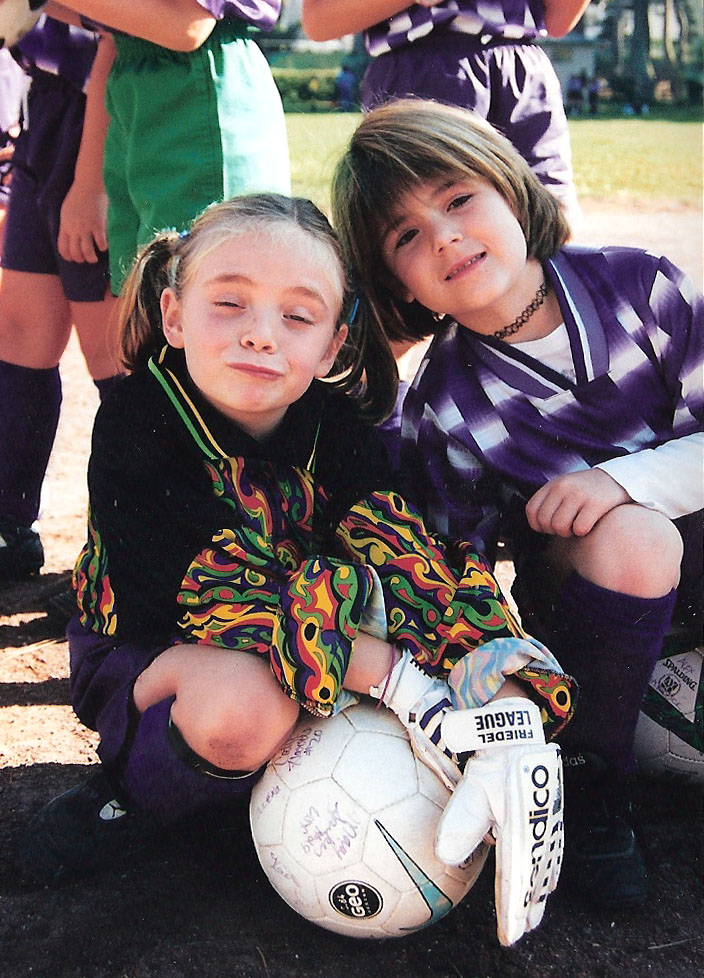 Two young girls kneel on the ground, dressed in soccer uniforms