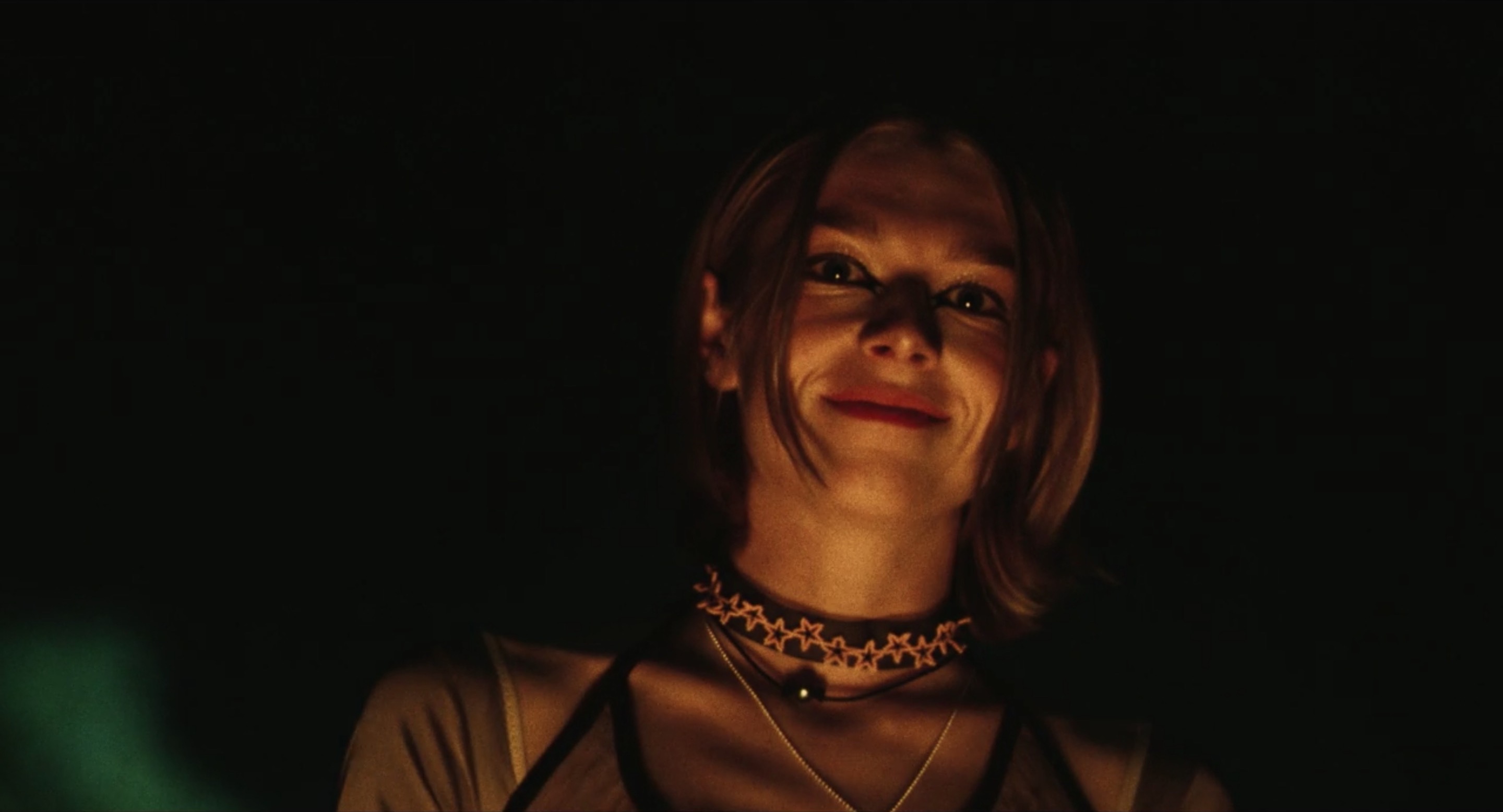 Hunter Schafer with dark black eyeliner is lit only by a fire. She has a short bob haircut.