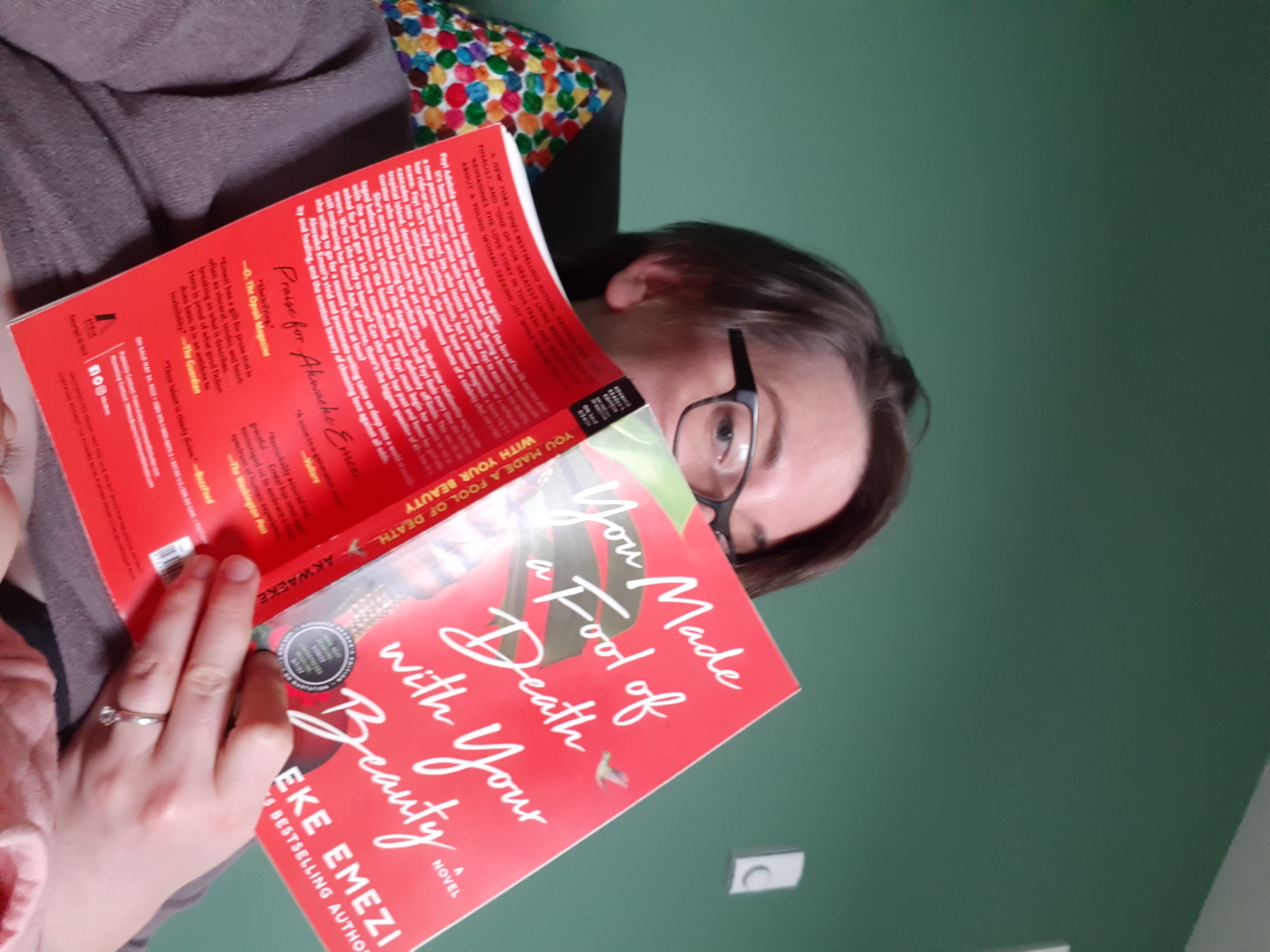 Casey looks over the top of a book with one eye. The book is "You Made a Fool of Death With Your Beauty." Casey is a white woman wearing a sweater with black glasses