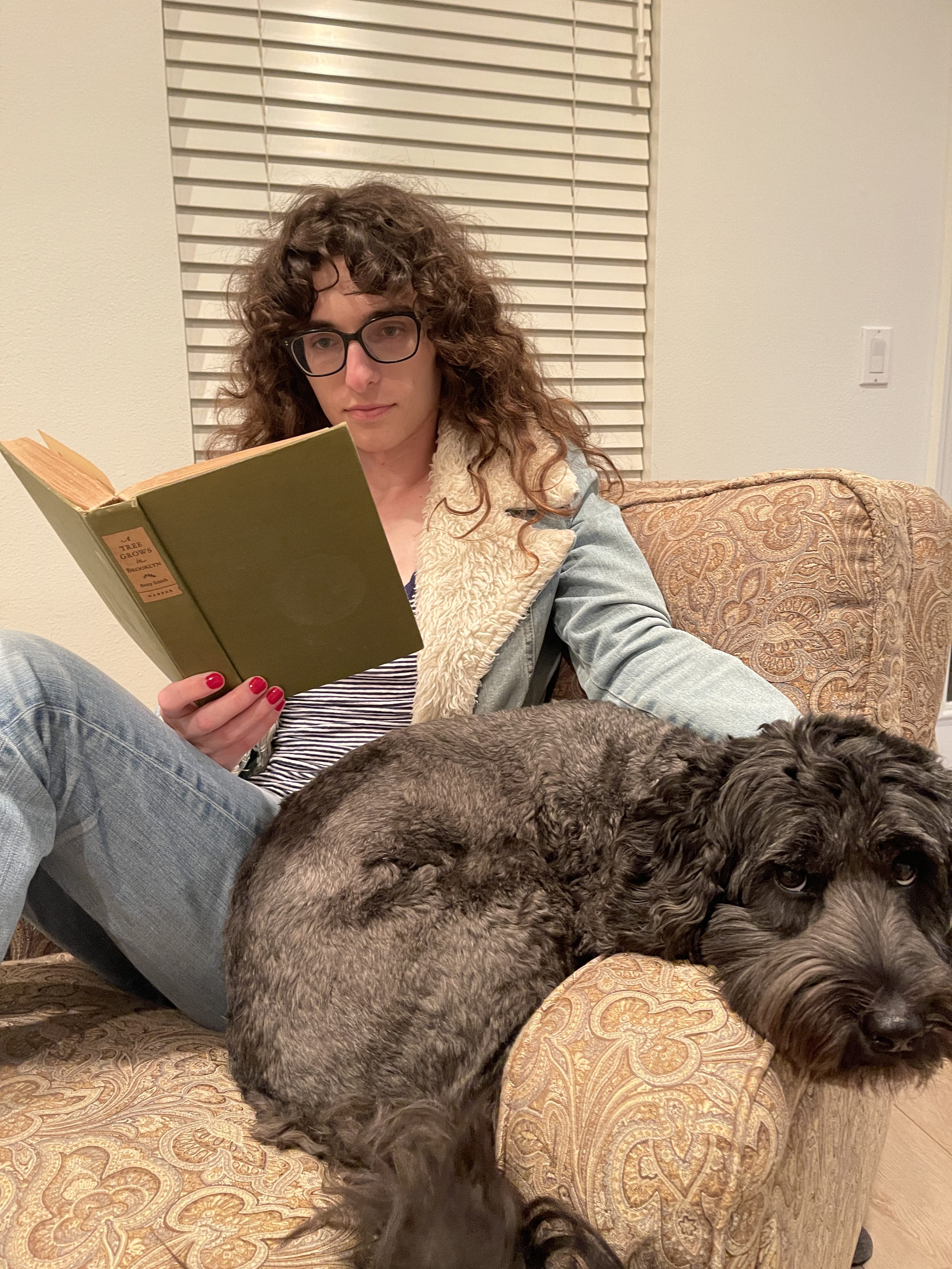 Drew reads from an old-looking tome on a comfy chair with a very adorable, curly-haired dog next to her. She has one hand on the dog and is holding the book with a hand with red nail polish. Drew is a white woman with curly hair, wearing glasses, a shearling lined jacket and jeans.