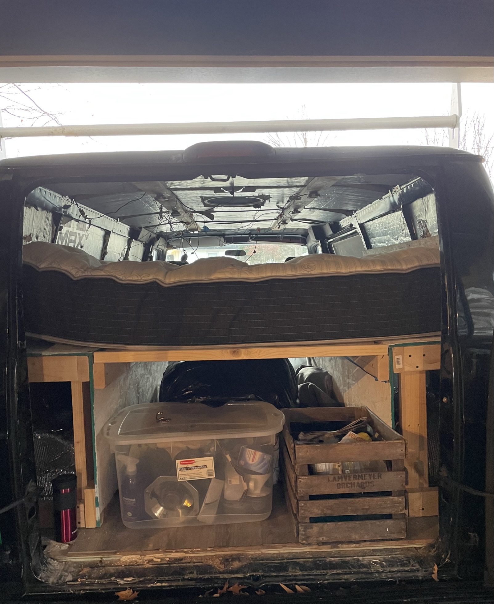 This is a look inside their van. There is a mattress on top of a wooden platform. Underneath are several storage crates and boxes holding supplies and items.