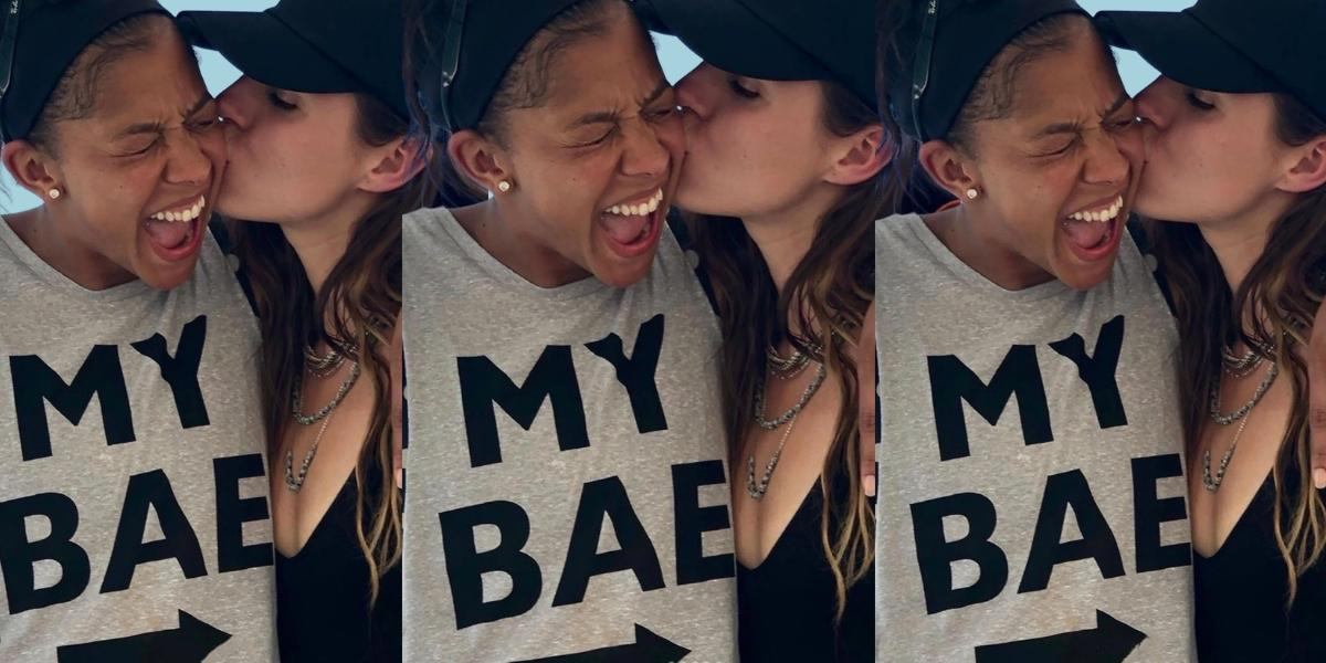 A cut out collage of Candace Parker and her wife. Her wife is kissing her cheek while Candace laughs. Candace is wearing a tank top that says 