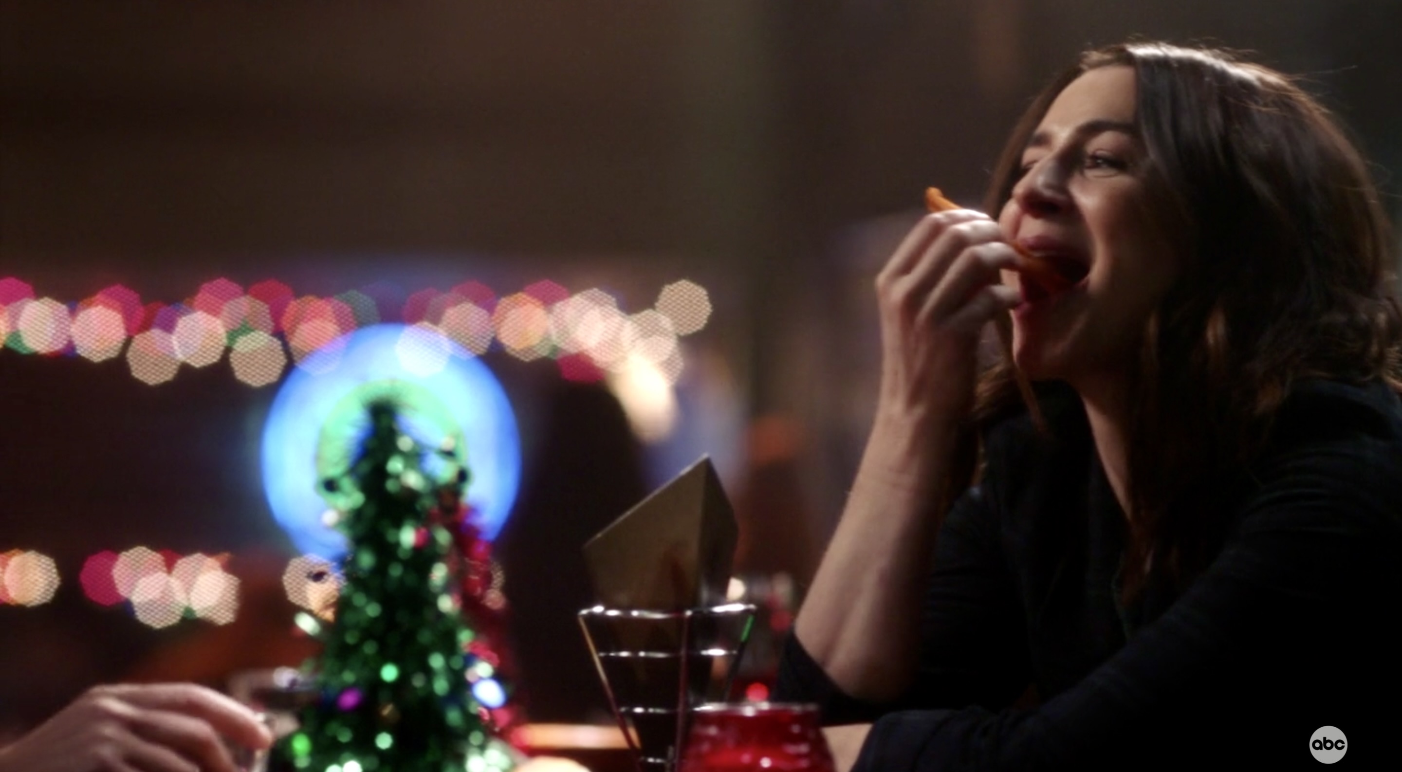 Amelia Shepherd smiles as she takes a big bite out of a sweet potato fry inside of Joe's Bar, with twinkly lights around her.
