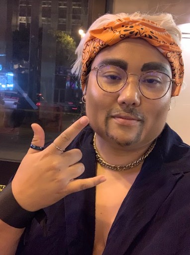Here, Em throws up the "I love you" sign with their hand. They have make-up on that thickens their eyebrows and gives them a beard. They are wearing a platinum wig and headband, chain, men's collared shirt, and thumb ring. They are inhabiting Guy Fieri!