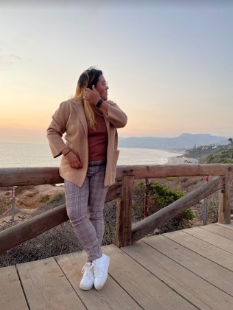Em is leaning against a fence in this picturesque scene, tucking her hair behind their ear. The sea extends out behind the beach behind Em. Em is wearing plaid pants and a camel blazer.