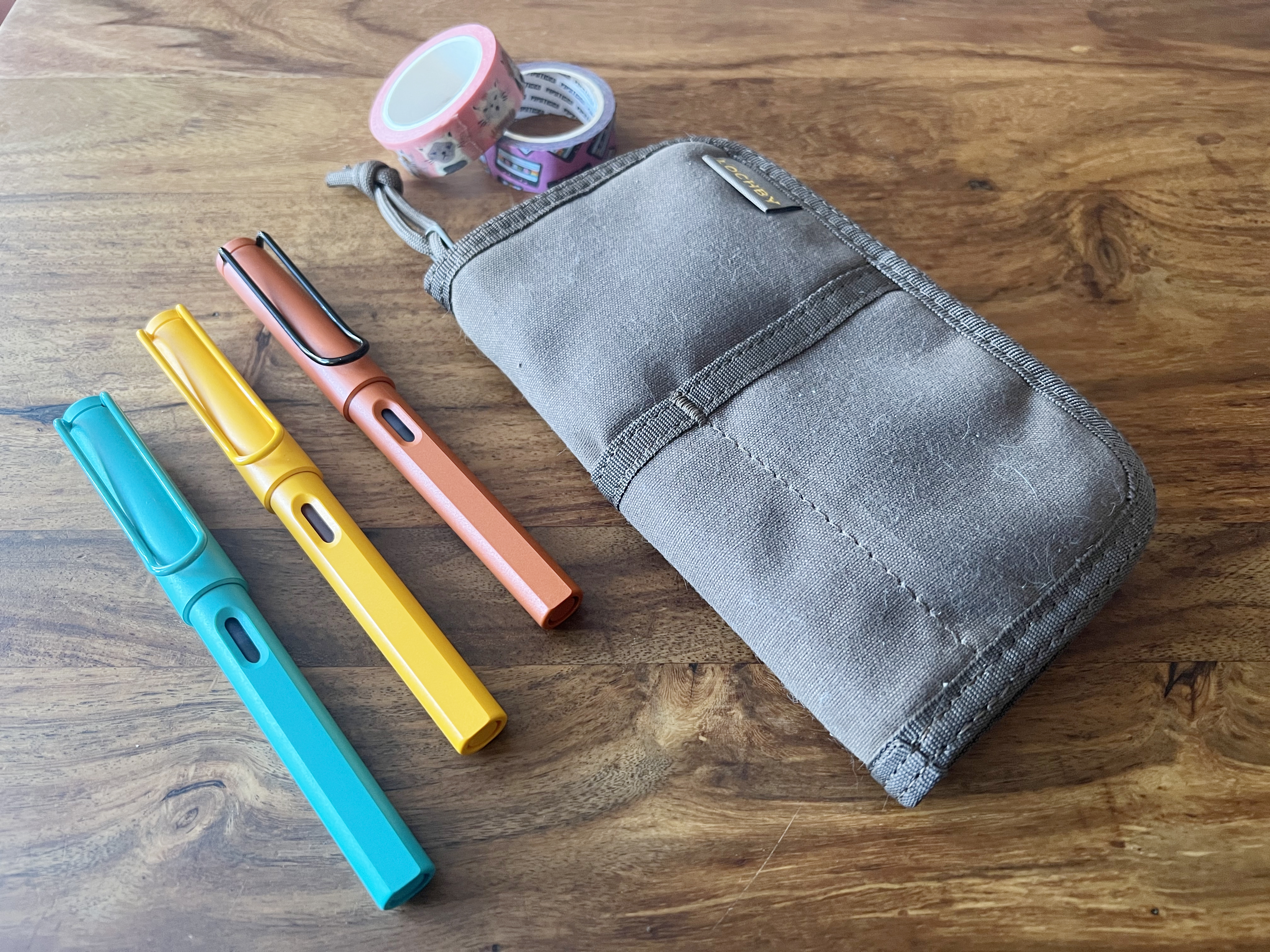 Three fountain pens rest on a wooden table near a carrying pouch and some washi tape.