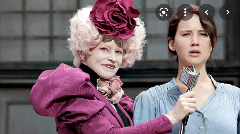 a screenshot from the hunger games movie with effie trinket, dressed super opulently, holding a microphone up to miserable looking jennifer lawrence