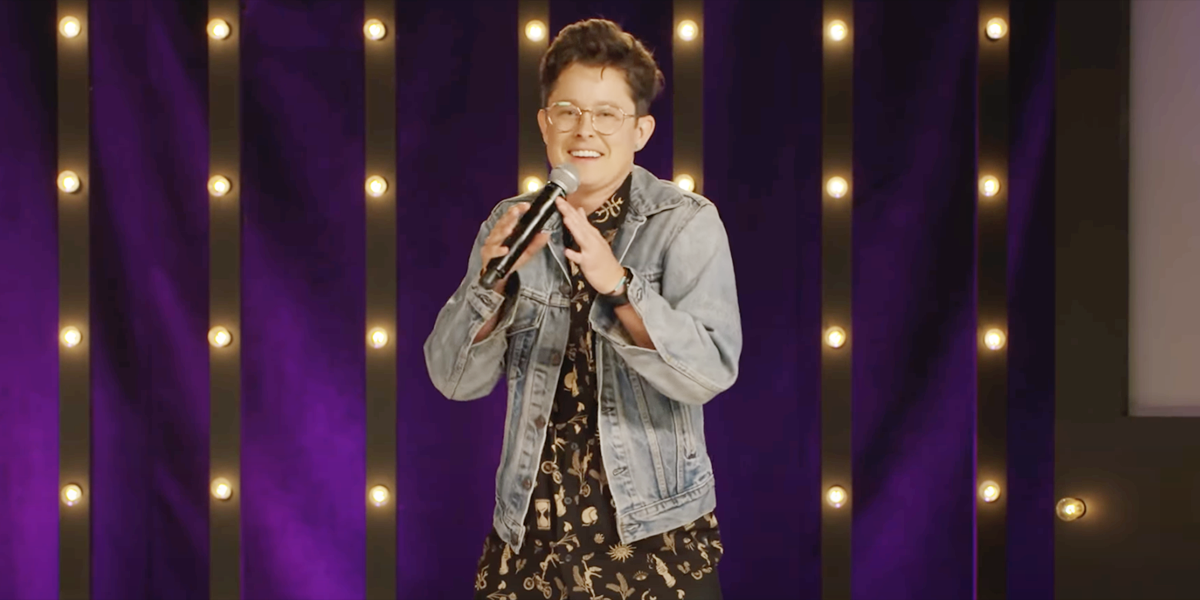 River Butcher in a jean jacket and black button-up standing on stage with a microphone laughing in front of a purple backdrop
