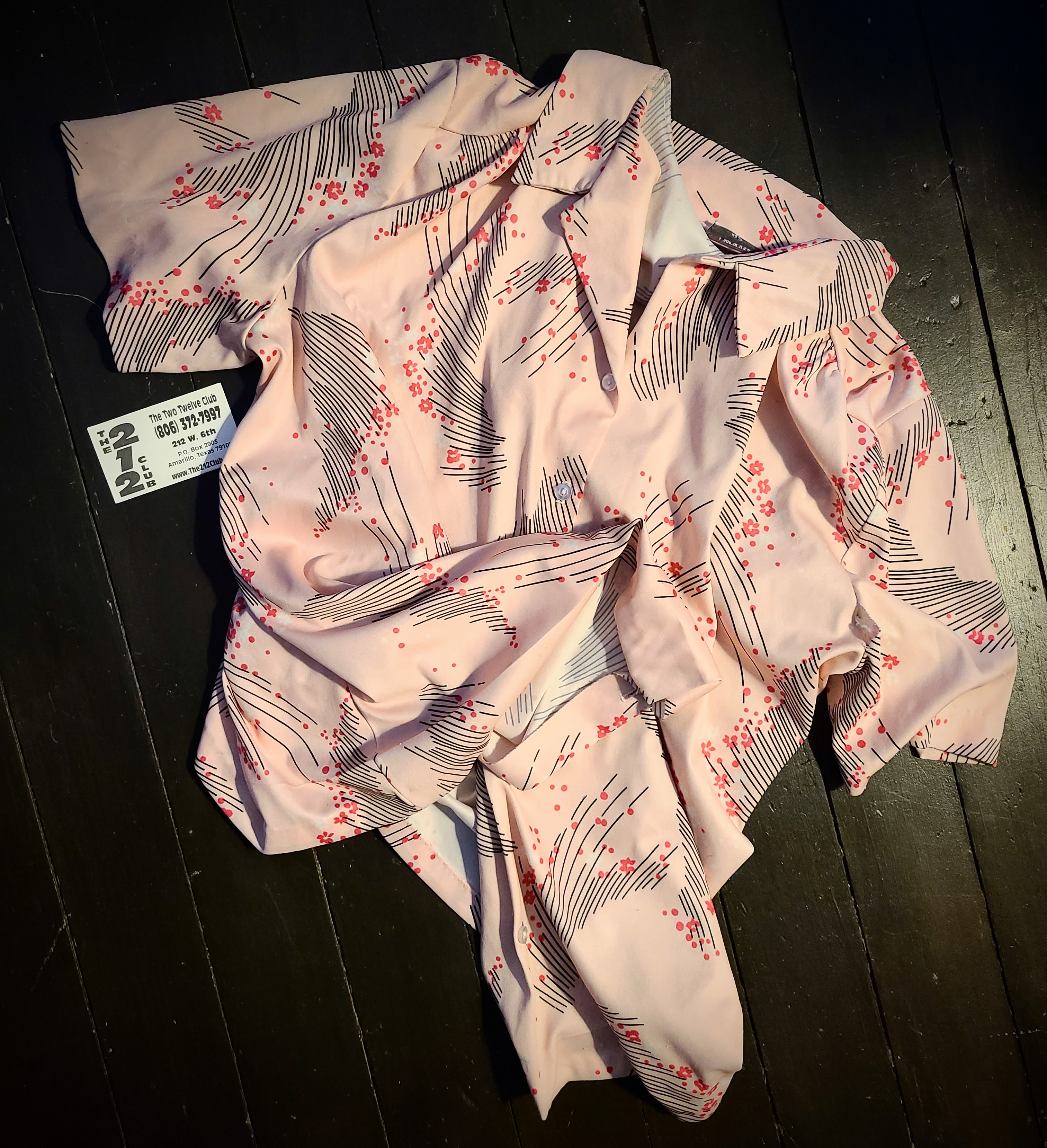 A polyester cool-toned pink short-sleeve button up top with an 80s or 90s looking black line and darker pink flower motif. The shirt is very loose and flowy which you can tell from the way it lies on the ground. There is a business card next to it for The 212 Club which is pronounced, according to the card, The Two Twelve Club. It's Amarillo address, 212 W. 6th ST. and phone number (806) 372-7997 are visible in the photo.