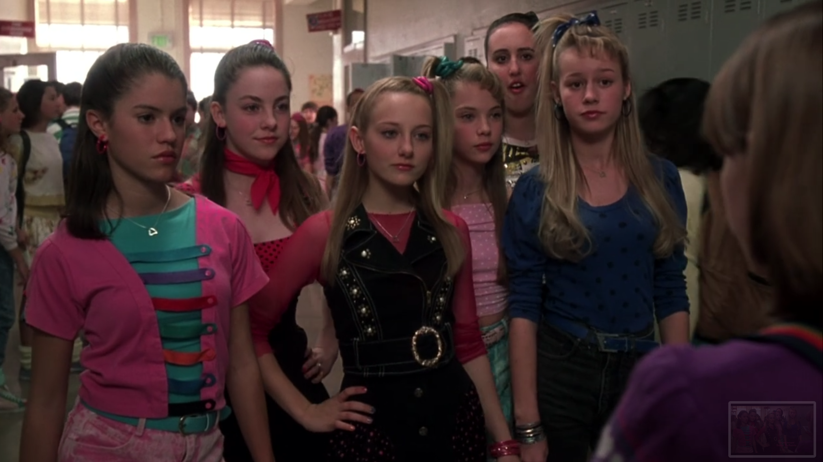 Six teen girls have ponytails and stand in a line in a school hallway. They are wearing very 80s looks.
