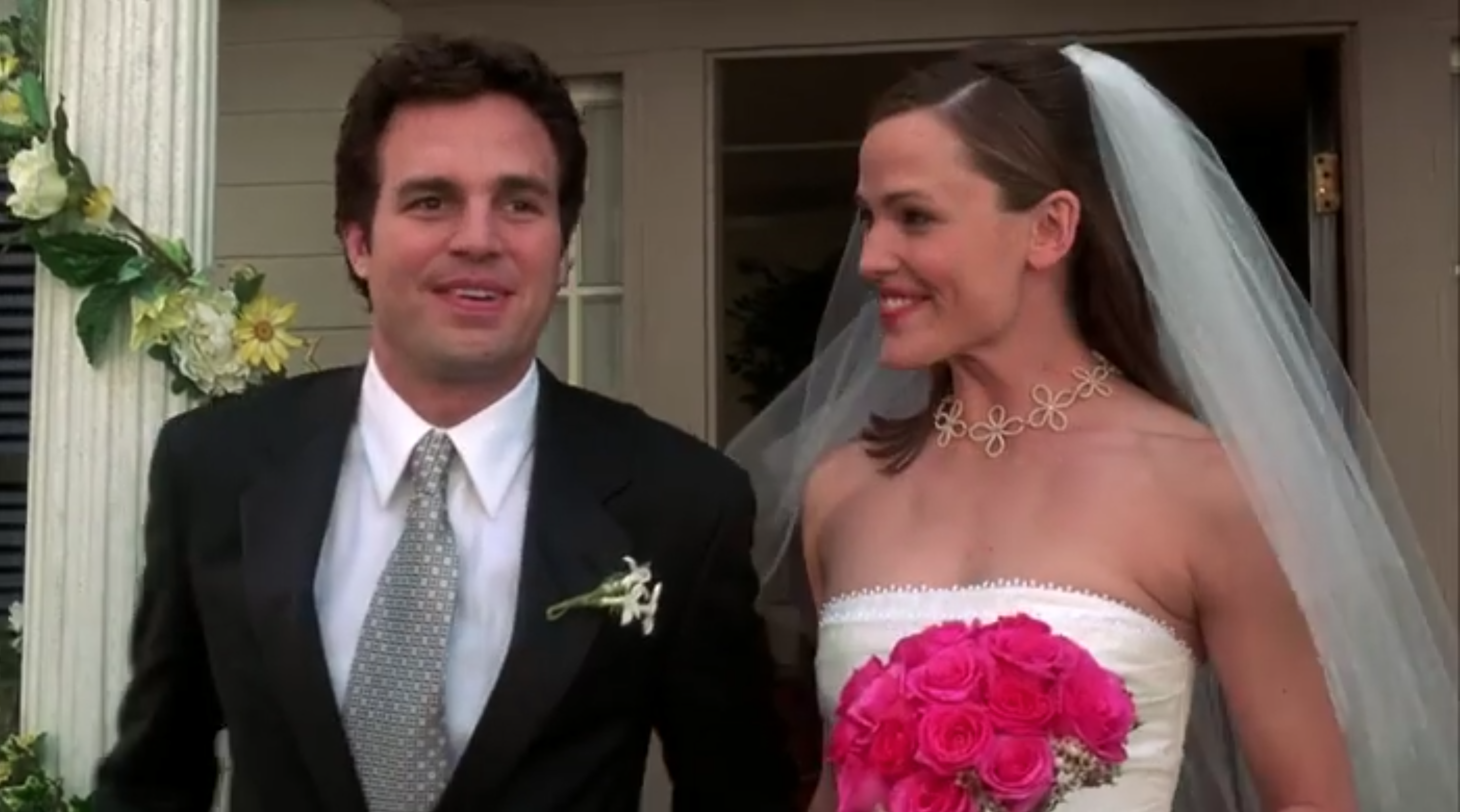 Matt is wearing a wedding suit, and Jenna is wearing a strapless wedding dress and holding a bouquet of pink flowers