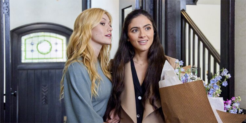 Arienne Mandi and Katherine McNamara stand in a doorway holding flowers in Hallmark's 