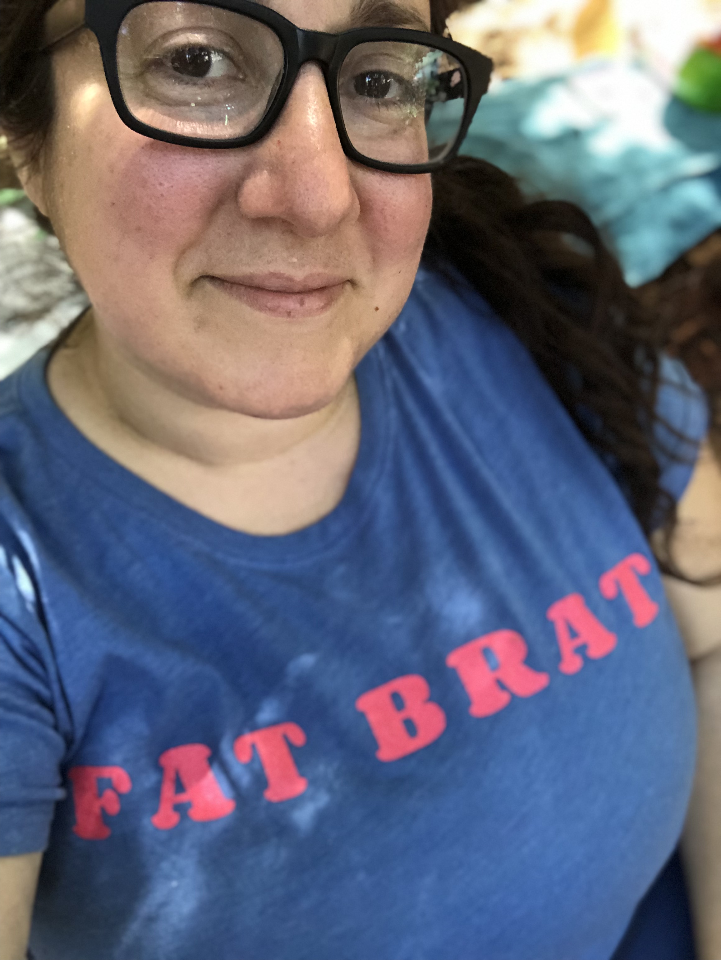 Vanessa, a white woman with long brown hair and glasses is wearing a shirt that reads "Fat Brat"
