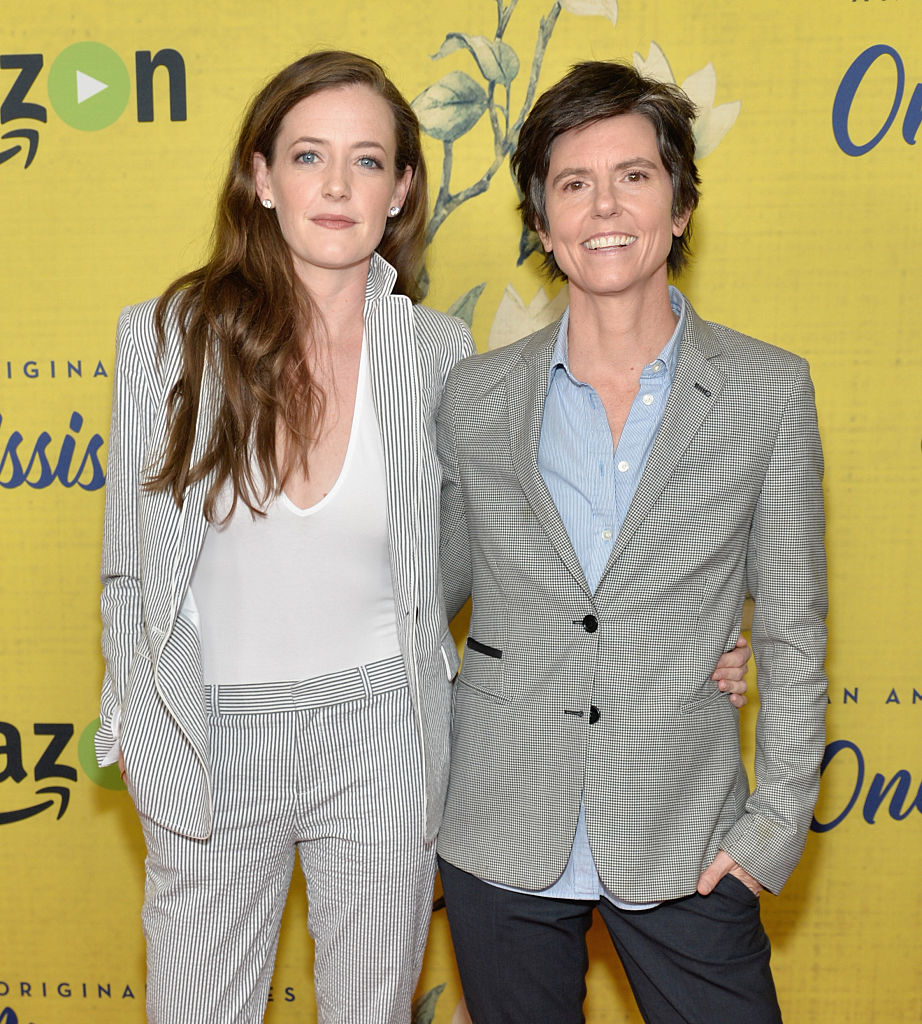 Stephanie Allynne and Tig Notaro attend the premiere of Amazon Instant Video's 'One Mississippi' at The London West Hollywood. Stephanie is wearing a white shirt with a striped blazer and matching pants. Tig is wearing a blue buttondown and a gray blazer. They have their arms around each other and are smiling at the camera.
