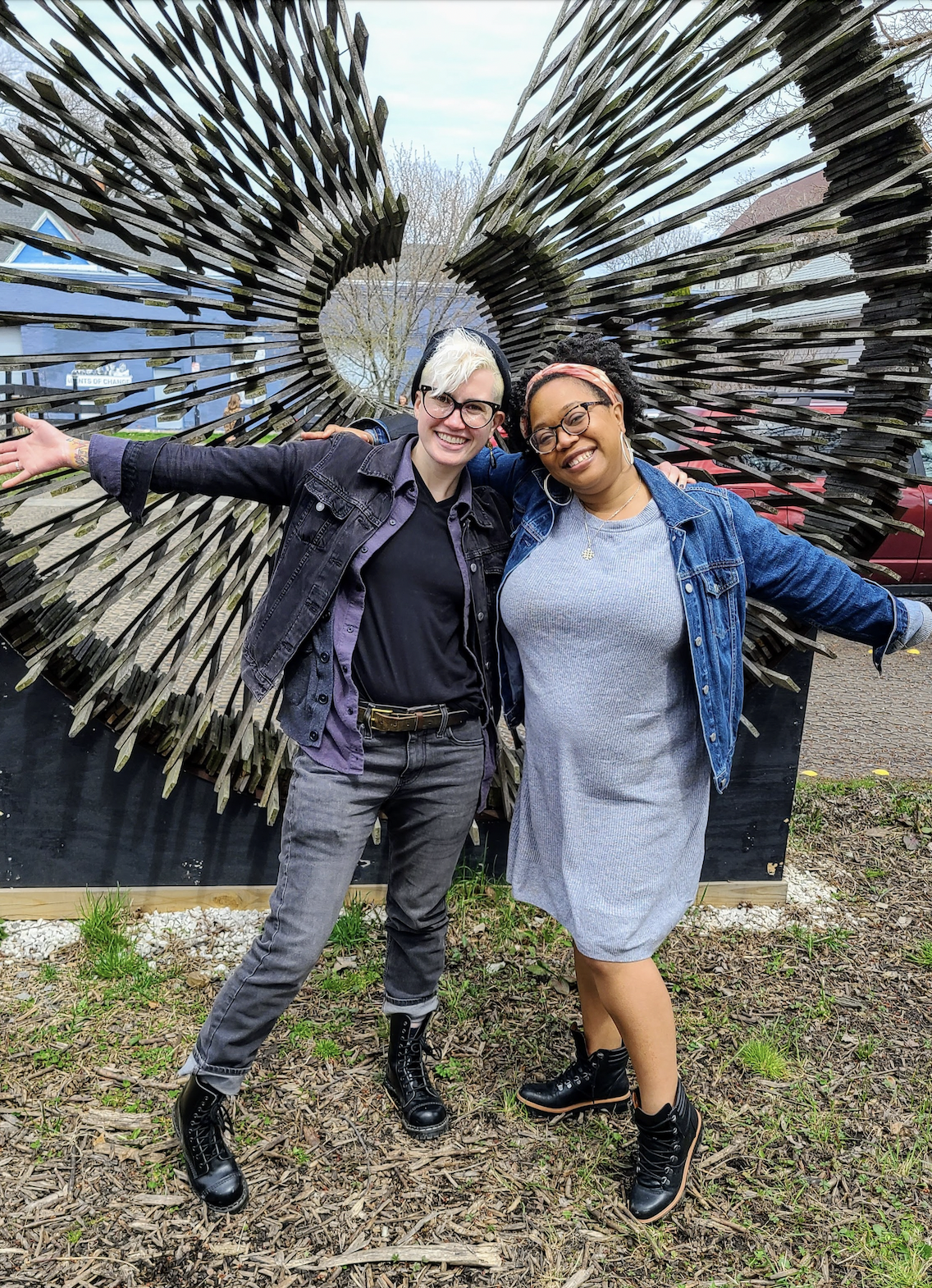 Nicole and Carmen pose arm in arm, with outside arms outsretched, smiling in front of a sculpture outdoors. Carmen is a Black woman who is wearing a headband tied in a bow at the top, and who has her curly hair parted in the center. She is wearing a jean jacket over a sweater dress and black boots that come up over the ankle as well as glasses and hoop earrings. Nicole is a white genderqueer human who is wearing a black jean jacket, gray jeans, a v neck black tee, a couple of random gray flannel layers and black combat boots along with a beanie and glasses. They are both smiling sweetly for the camera!
