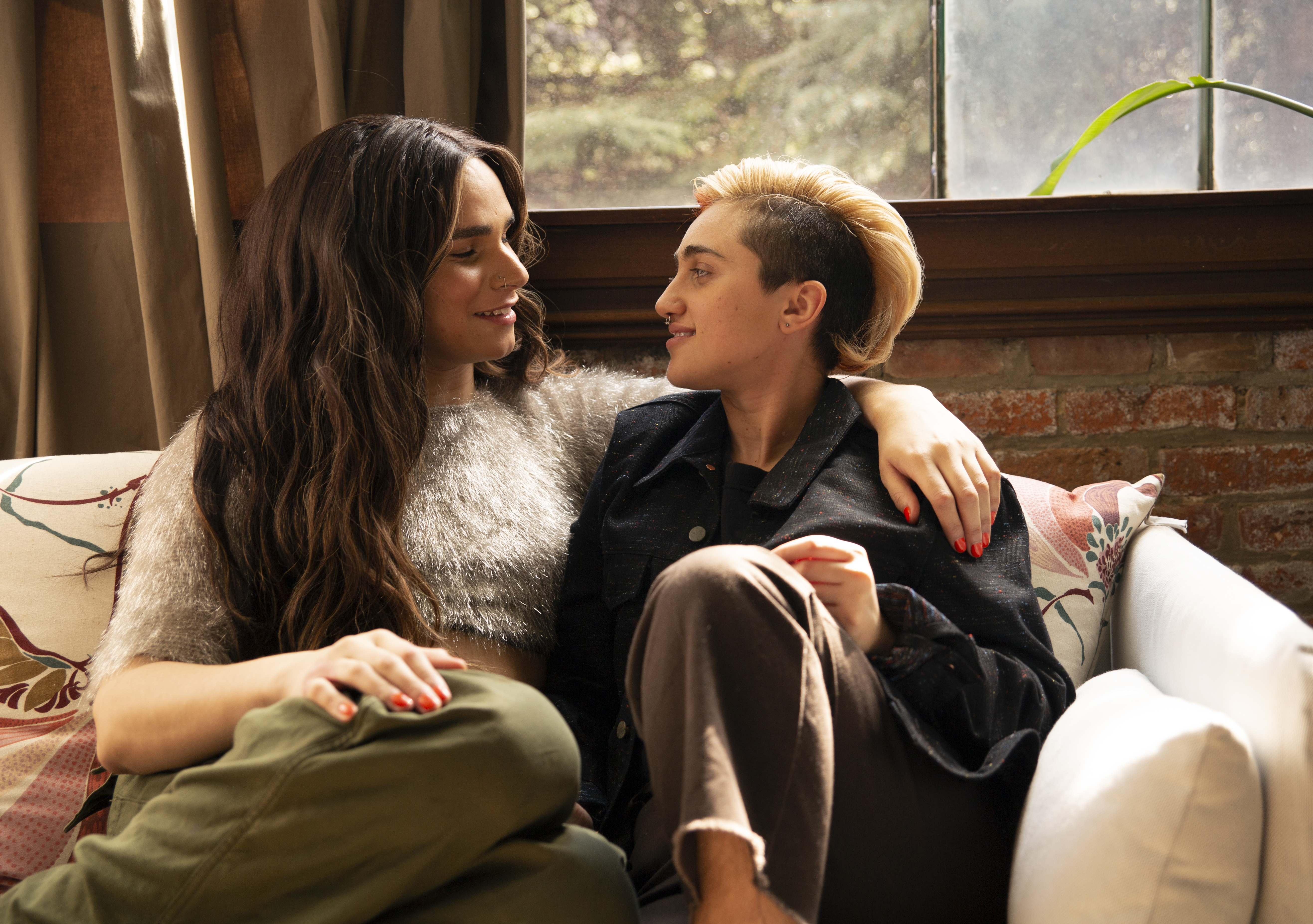 A transfeminine non-binary person and transmasculine gender-nonconforming person look at each other on a couch