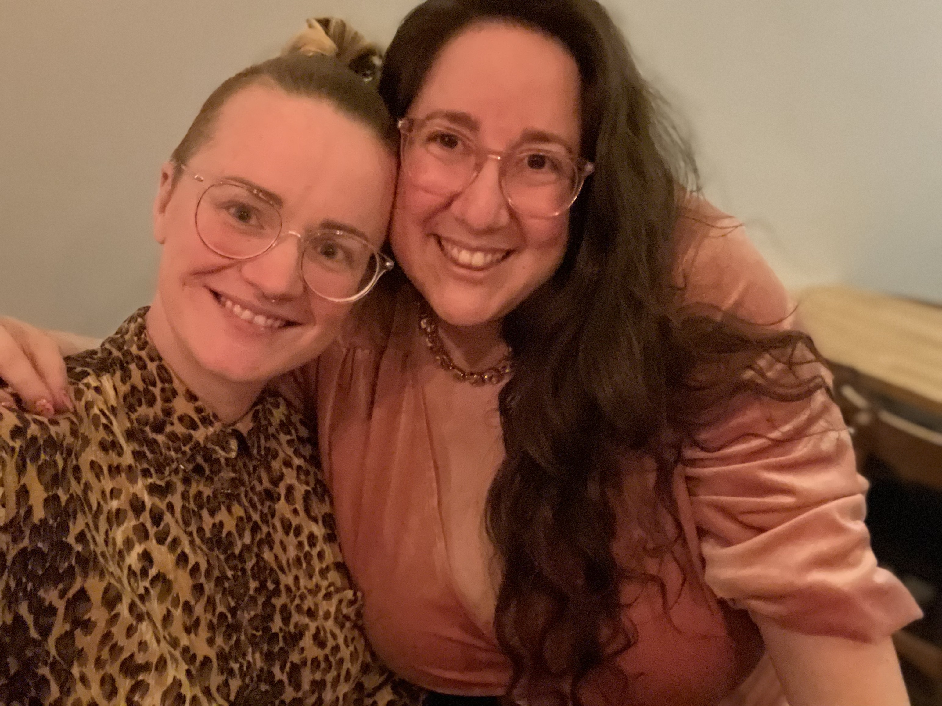 PB and Vanessa pose for a photo while smiling. PB is wearing glasses and a cheetah print button up. Vanessa is wearing a pink velvet dress and necklace. PB is white and has blonde hair that is long on top and up in a bun with shaved sides. Vanessa is also white with long brown hair she is wearing down.
