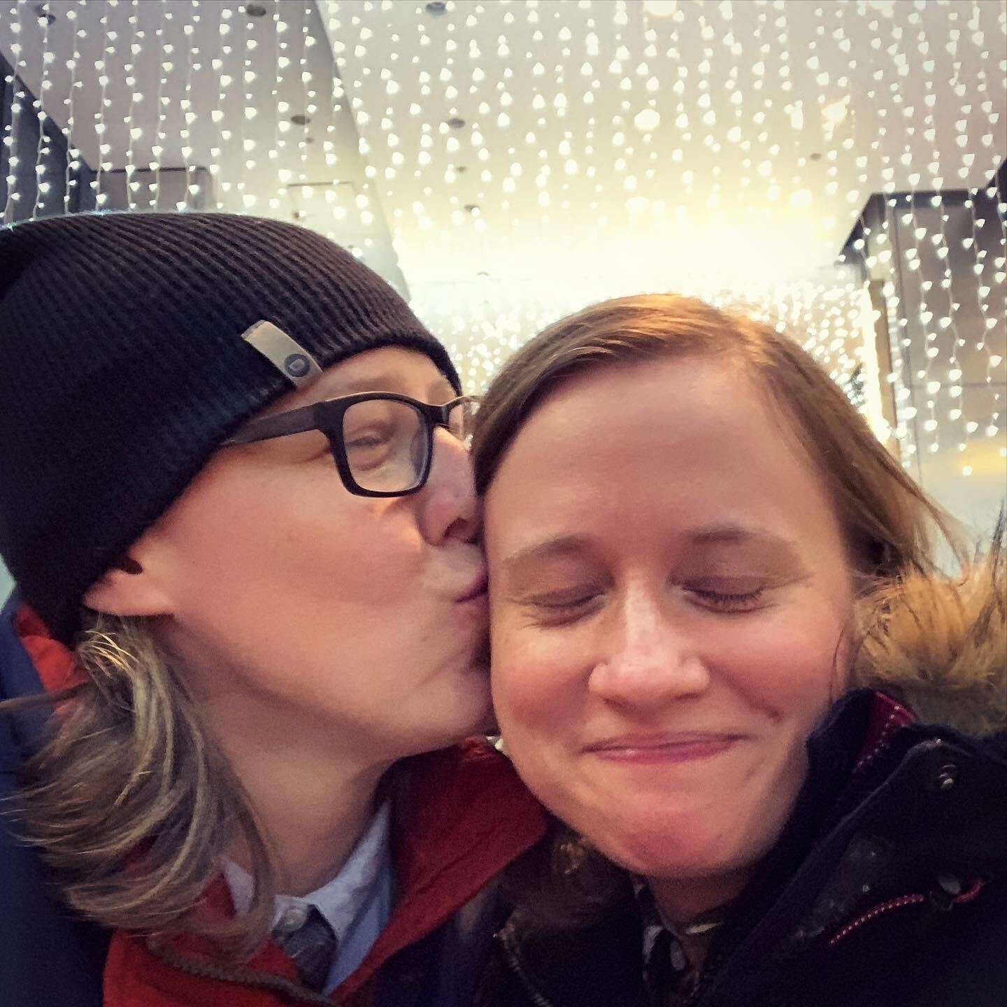 Heather and Stacy are pictured in this photo. In the photo, Heather, a soft butch white woman wearing black rimmed glasses and a beanie, kisses Stacy, a white woman with light brown hair, on the cheek. They are both smiling and both look happy. You can see string lights glowing behind them..