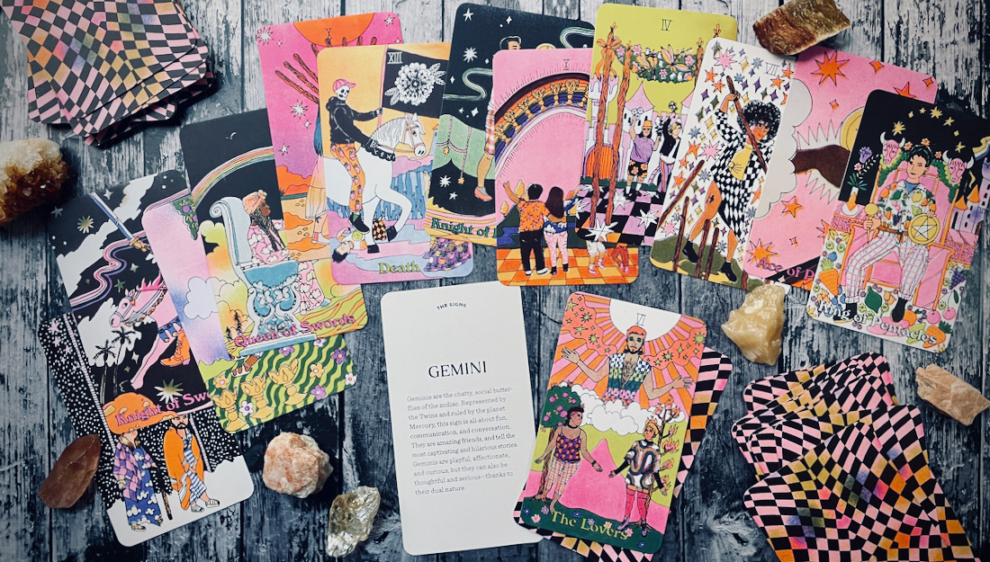 A collage of tarot cards on a grey wood table, the cards are in shades of Pink and spring warm hues. In the middle is a card that says Gemini in script font.