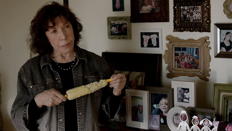 Elle eats a corn on the cob while standing in front of a display of family photos and childhood crafts