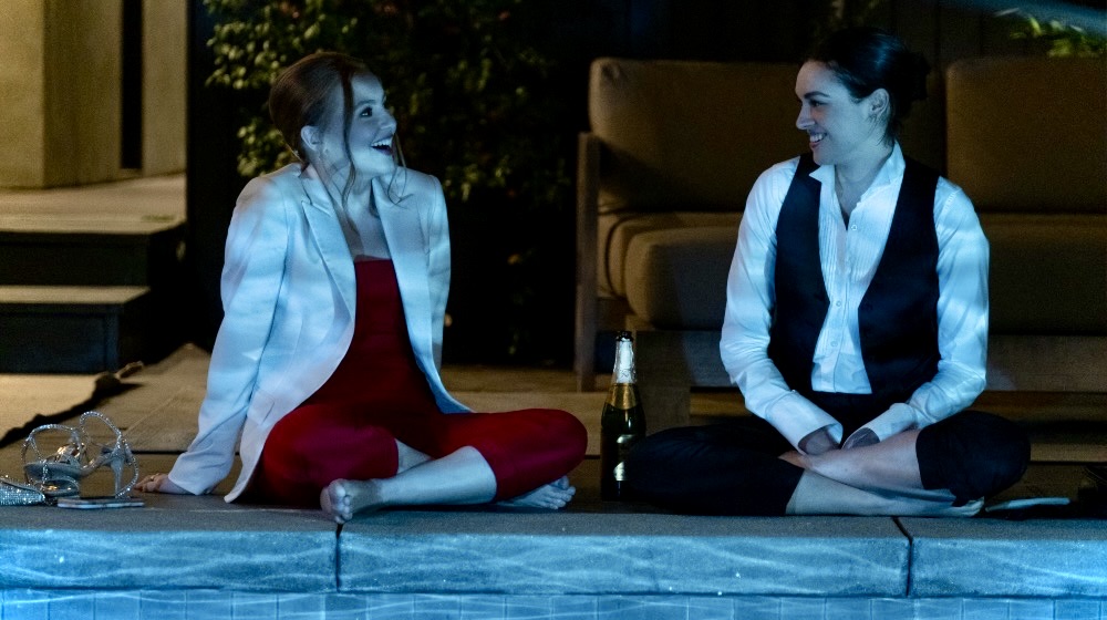 In Love Victor Season 3, Lake is in a red jumpsuit and white suit coat, sitting cross legged at a pool next to Lucy, who is in a cater waiter uniform that is unbuttoned.