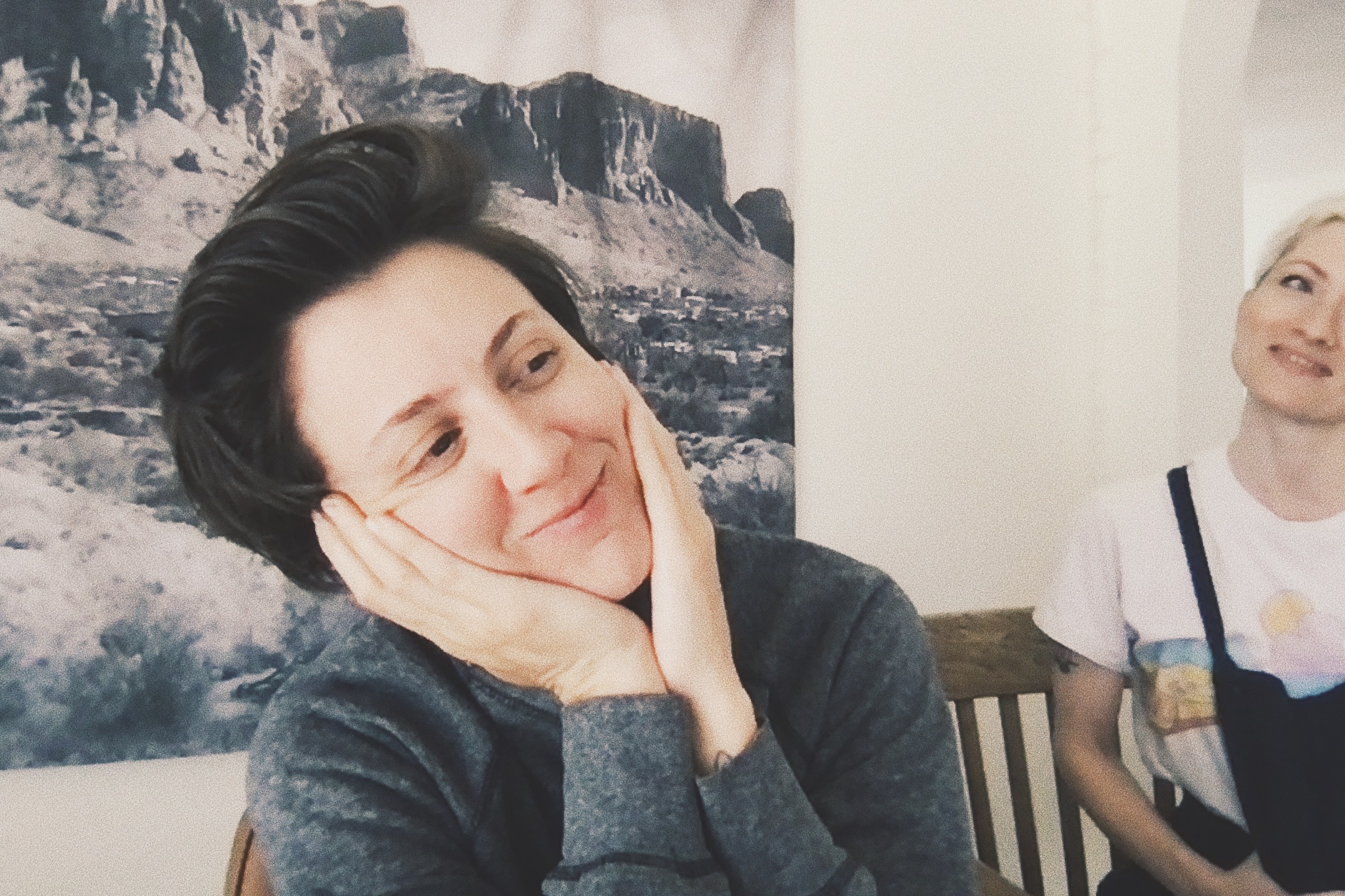 A photo of Amanda and Laneia sitting at the table in Laneia's house. Amanda looks like she is laughing and has her face in her hands. Amanda (she/her) is a white, androgynous 38 year old person with a dark brown shaggy haircut Laneia (she/her) is a short white 41 year old woman with a blonde pixie cut Amanda is seen here wearing a sweater. Laneia is wearing overalls over a tee shirt. 