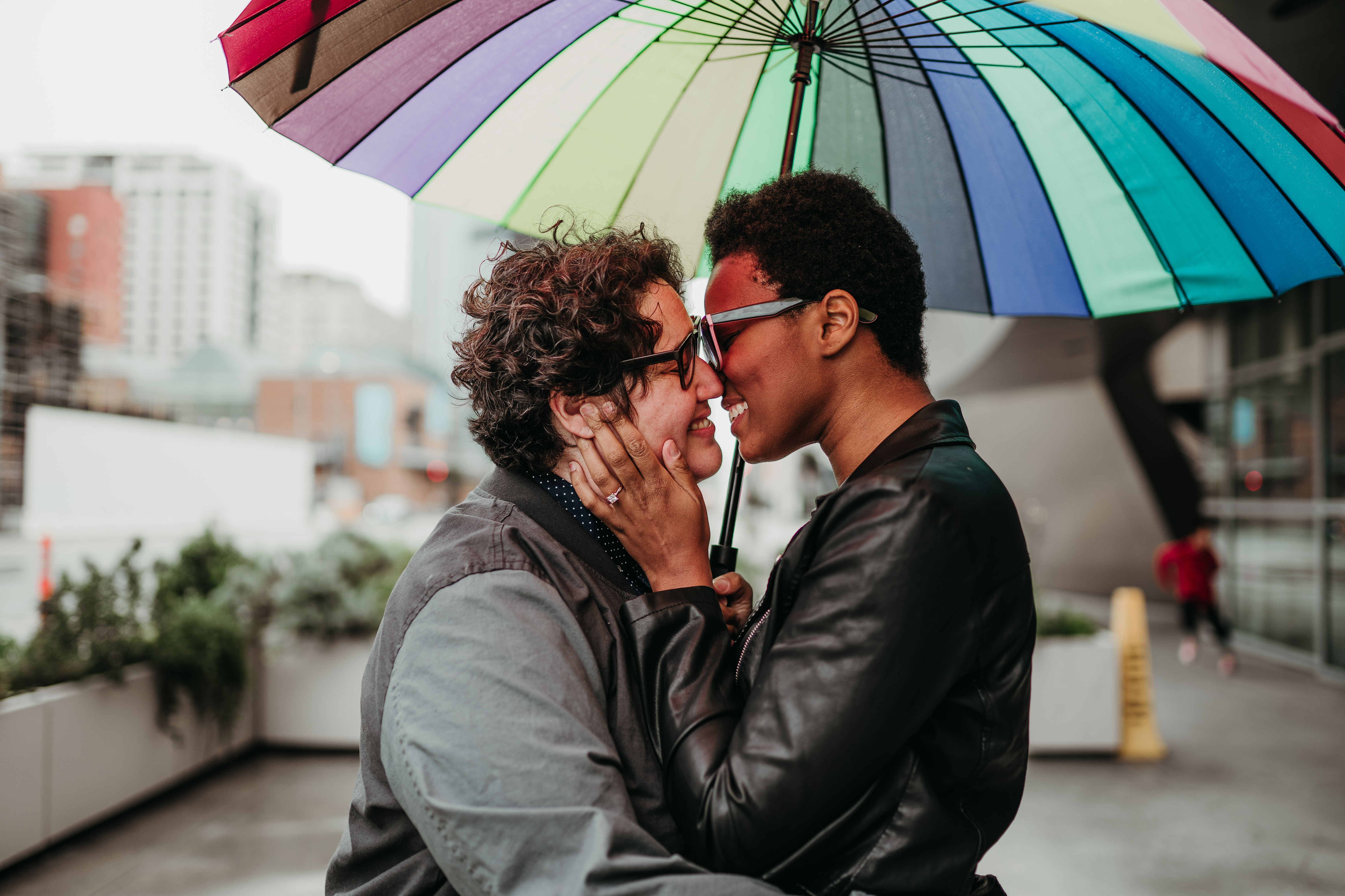 Sa'iyda clasps her fiancee's face in her hands and looks at her lovingly. They stand beneath a rainbow umbrella. Sa'iyda is a Black woman who is wearing blue glasses and a leather jacket and has short hair. Beth, her fiancee, is a white woman with curly short brown hair and black glasses who is wearing a gray jean jacket. An engagement ring is conspicuously visibile on Sa'iyda's hand!