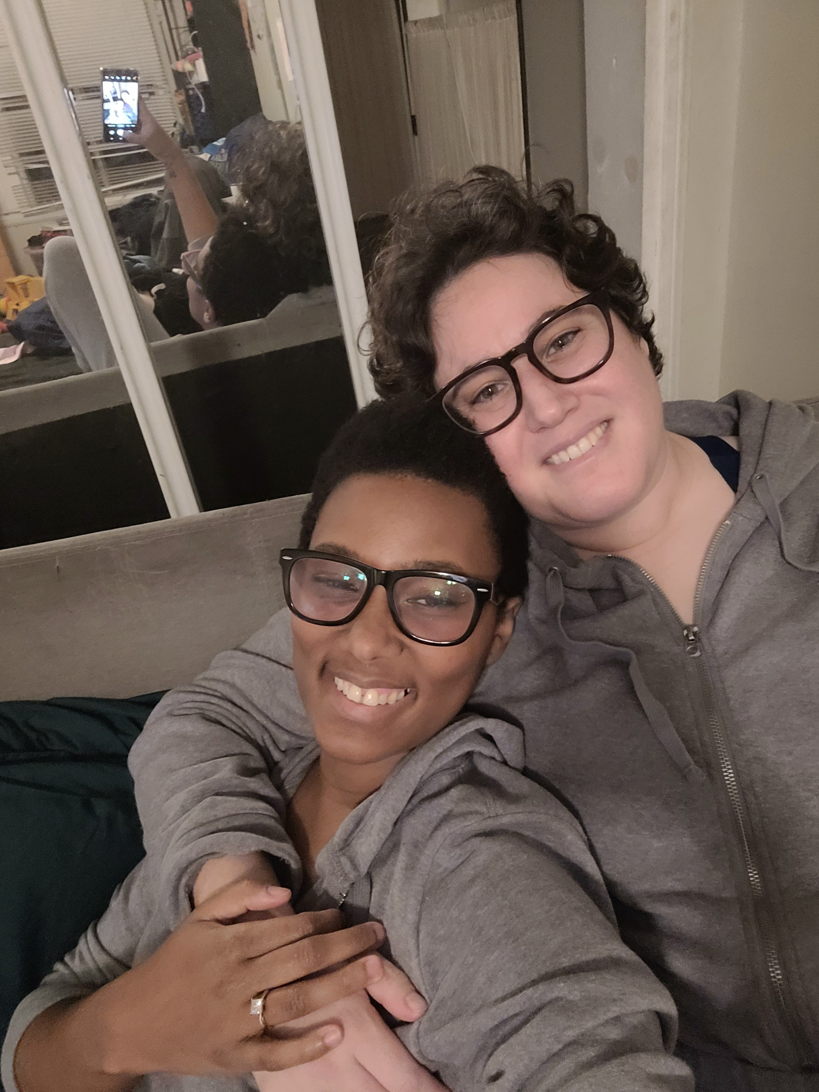 Sa'iyda and Beth cuddle while wearing matching gray hoodies on a couch. They're both smiling.