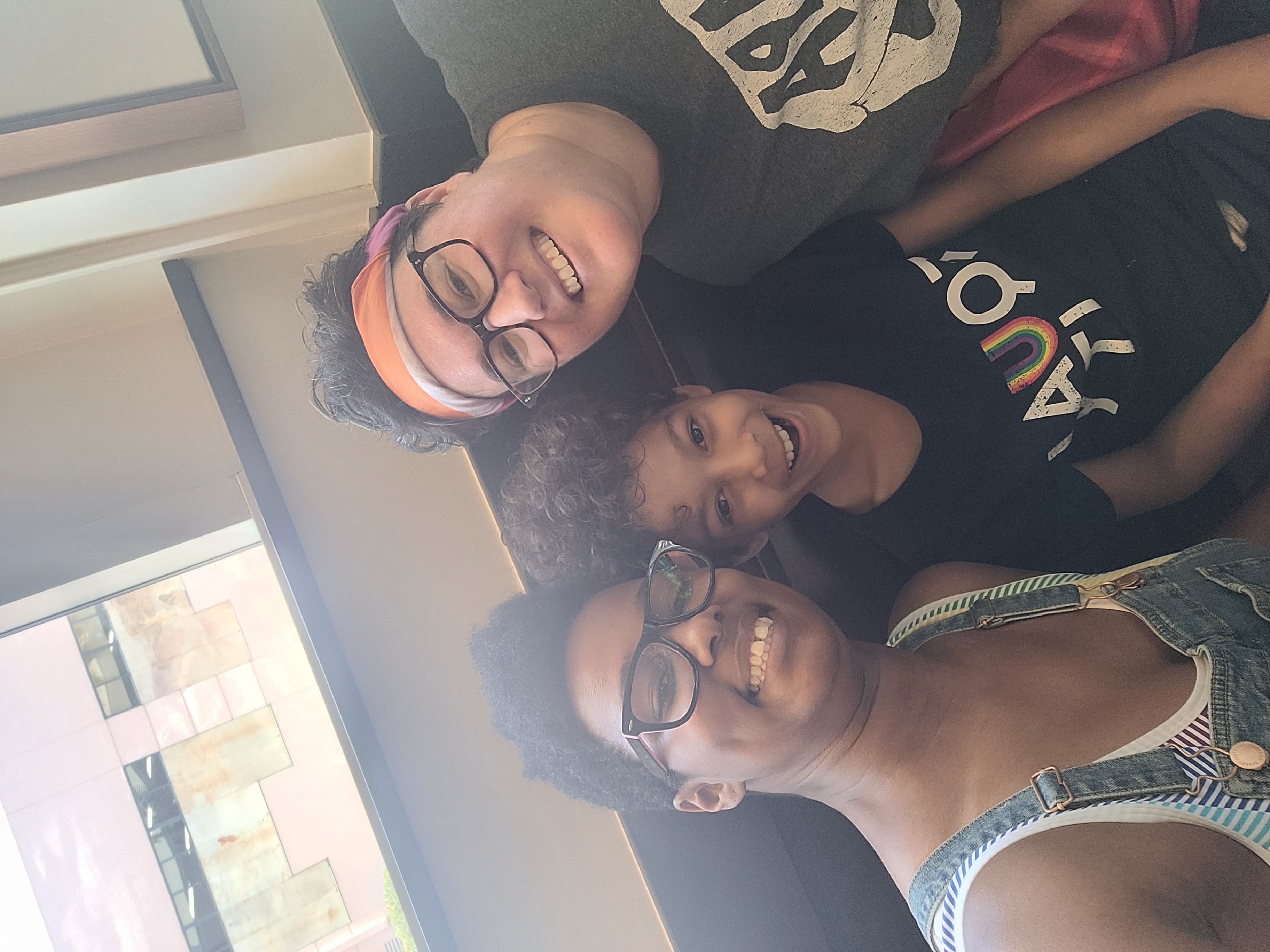 Sai, her fiancee Beth and her kid. Sai is a Black woman with short hair and glasses wearing a tank top. Beth is a white woman with short brown hair and glasses.