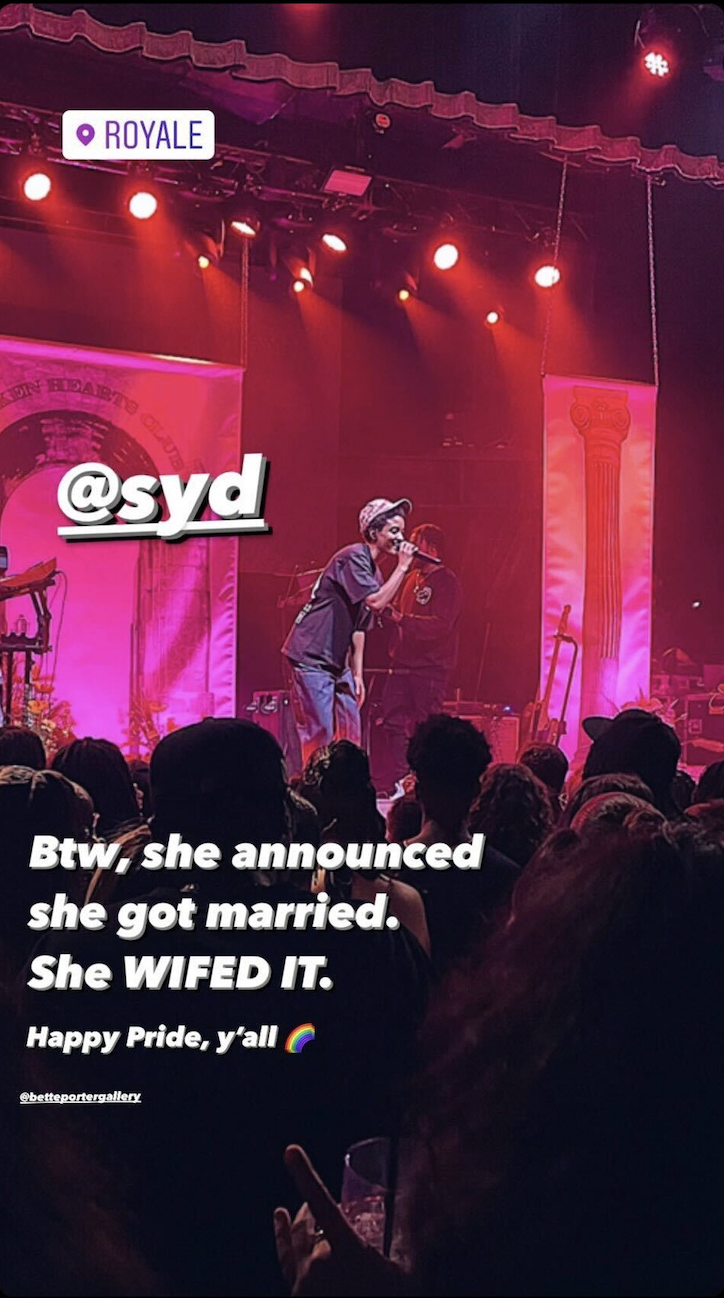 A vertical screenshot of Sad at a concert in Boston, she is on stage in front of a large crowd with red lights behind her. Superimposed on the image is the following test "Syd" / "Btw, she announced she got married. She WIFED it. Happy pride, y'all"
