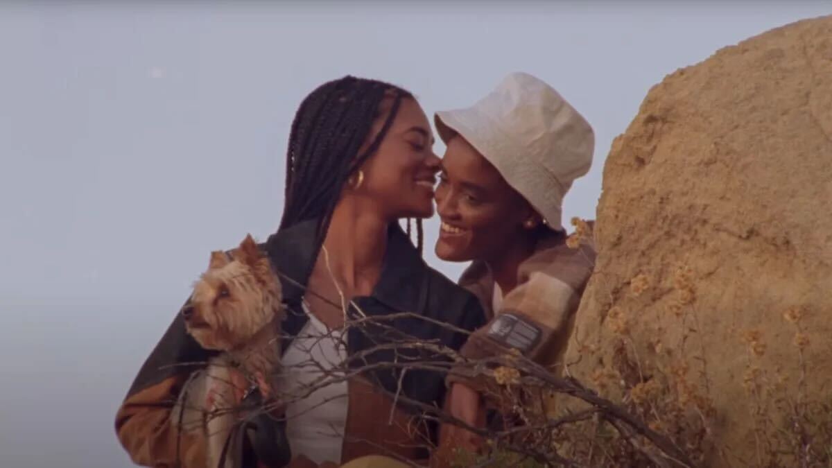 Syd and Ariana Simone, who just got married, together in the music video for Fast Car, Ariana Simone is holding a fluffy small dog, has on a denim jacket, and braids past her shoulders. Syd has on a khaki bucket cap. They are leaning next to a rock as tall as them and a blue sky behind them.