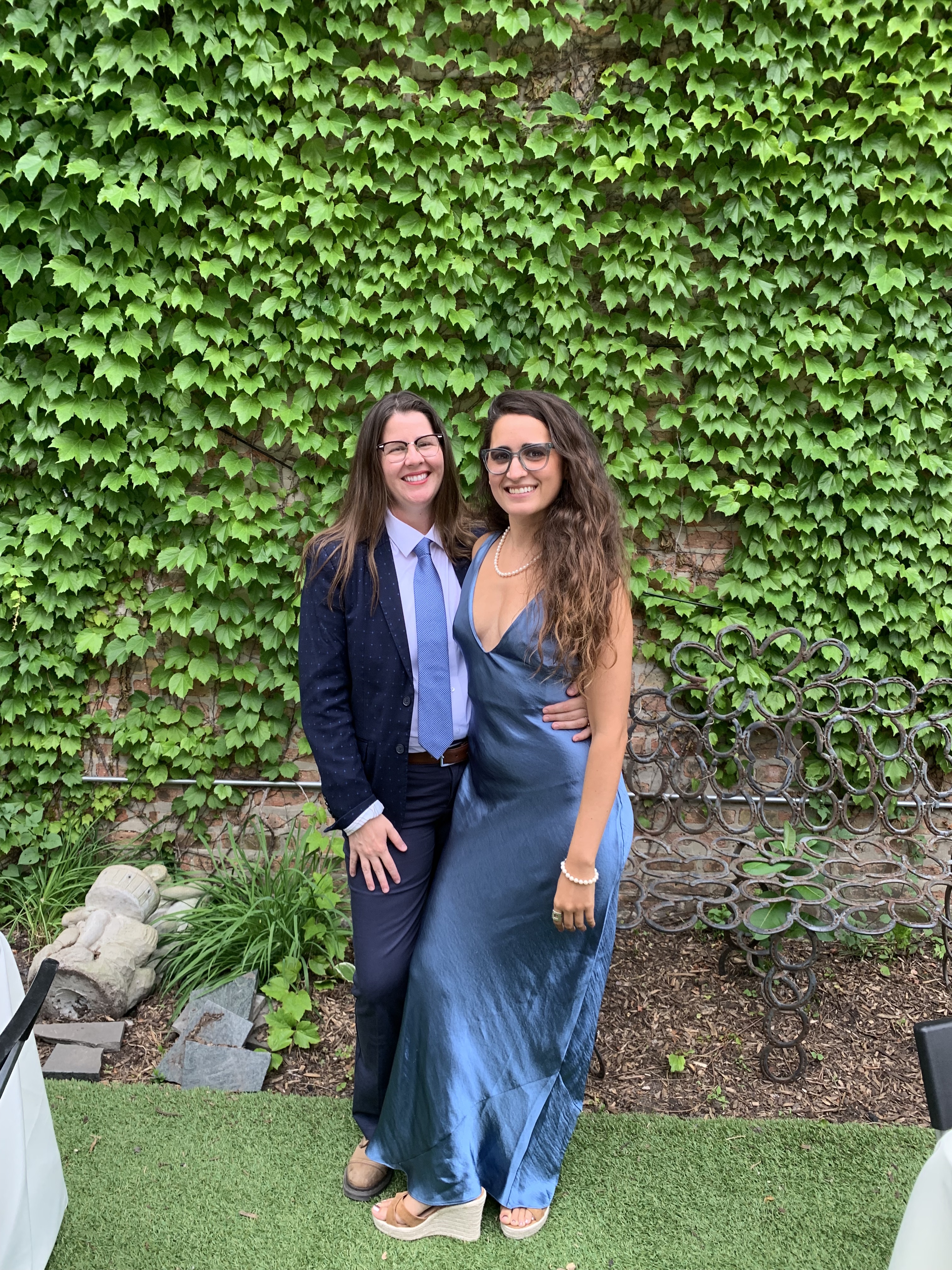 Kayla stands with her girlfriend Kristen in front of an ivy covered wall. Kristen is a white butch human wearing a blue suit. Kayla is a South Asian woman wearing a satiny blue dress with long brown hair and glasses.