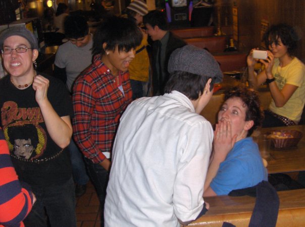 Peter, a white person with a septum piercing wearing glasses, a black David Bowie shirt and a newsboy cap smiles and dances; while Khlaeb, a Black person with straight, shoulder length black hair, wears a red flannel shirt, and Kate, a white person with short, black hair, wears a newsboy cap and a white button-up shirt. Khaleb and Kate lean over a booth a gossip with Mica, a white person with short, curly brown hair, who wears a blue shirt and has her hands over her mouth. In the background, Pidgeon, who has brown skin and shoulder-length black hair, wears a yellow T-shirt and takes a photo on a small, silver phone.