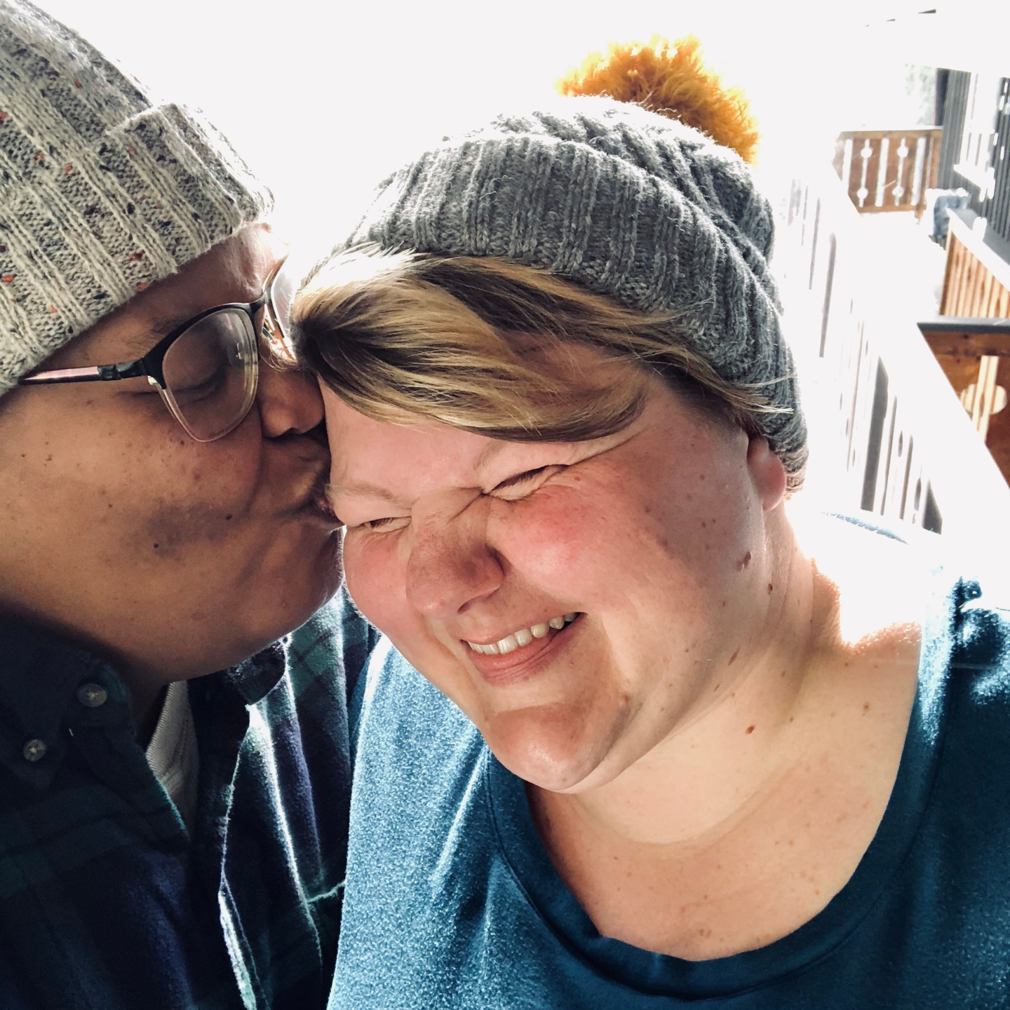 a close up photo of both shea and Jane in knit hats, smiling. shea kisses Jane on the cheek.