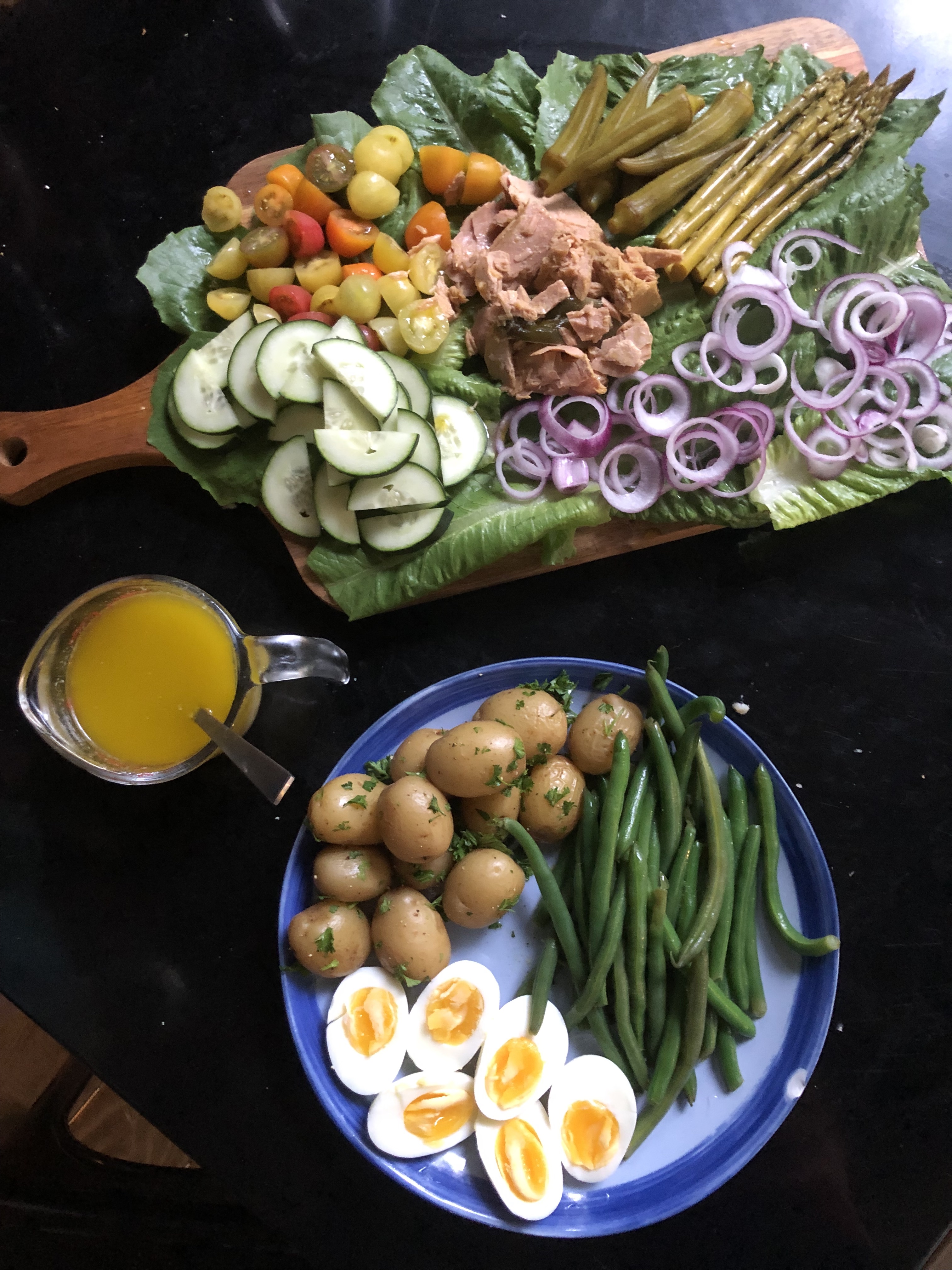 A tuna nicoise on a wood board and blue plate. On the blue plate, there are potatoes dressed in parsley, medium boiled eggs, and green beans. On the wood board, there's romaine lettuce, raw red onion, sliced cucumber, sliced multicolor cherry tomatoes, pickled asparagus, pickled okra, and a pile of tuna with jalapeño. There's also a small measuring cup full of dressing.