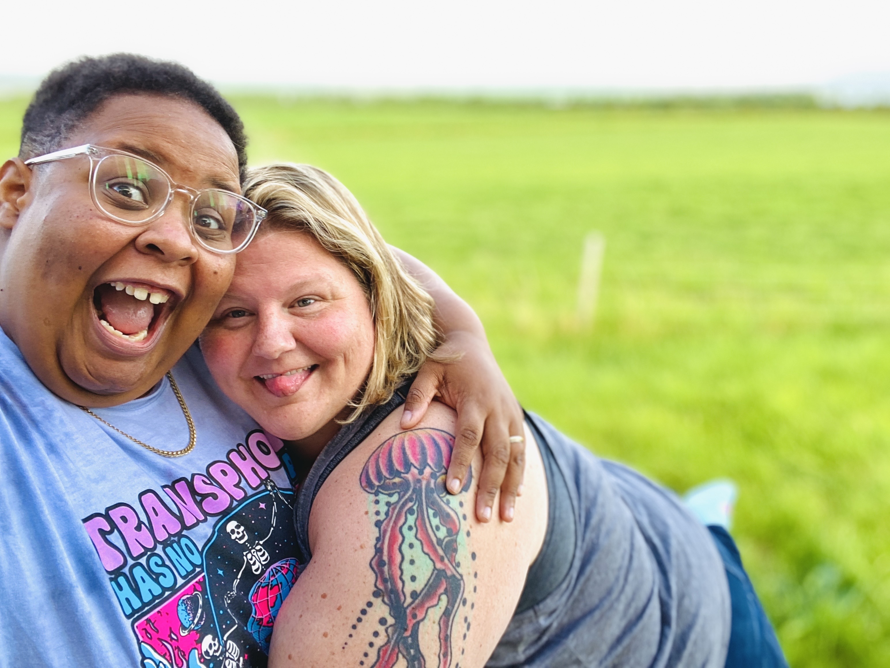 shea and their wife Jane are holding each other and smiling hugely on a field of bright green grass. shea is a Black nonbinary human with short hair and glasses. Jane is a white woman with a jellyfish tattoo on her arm and blonde shoulder length hair.