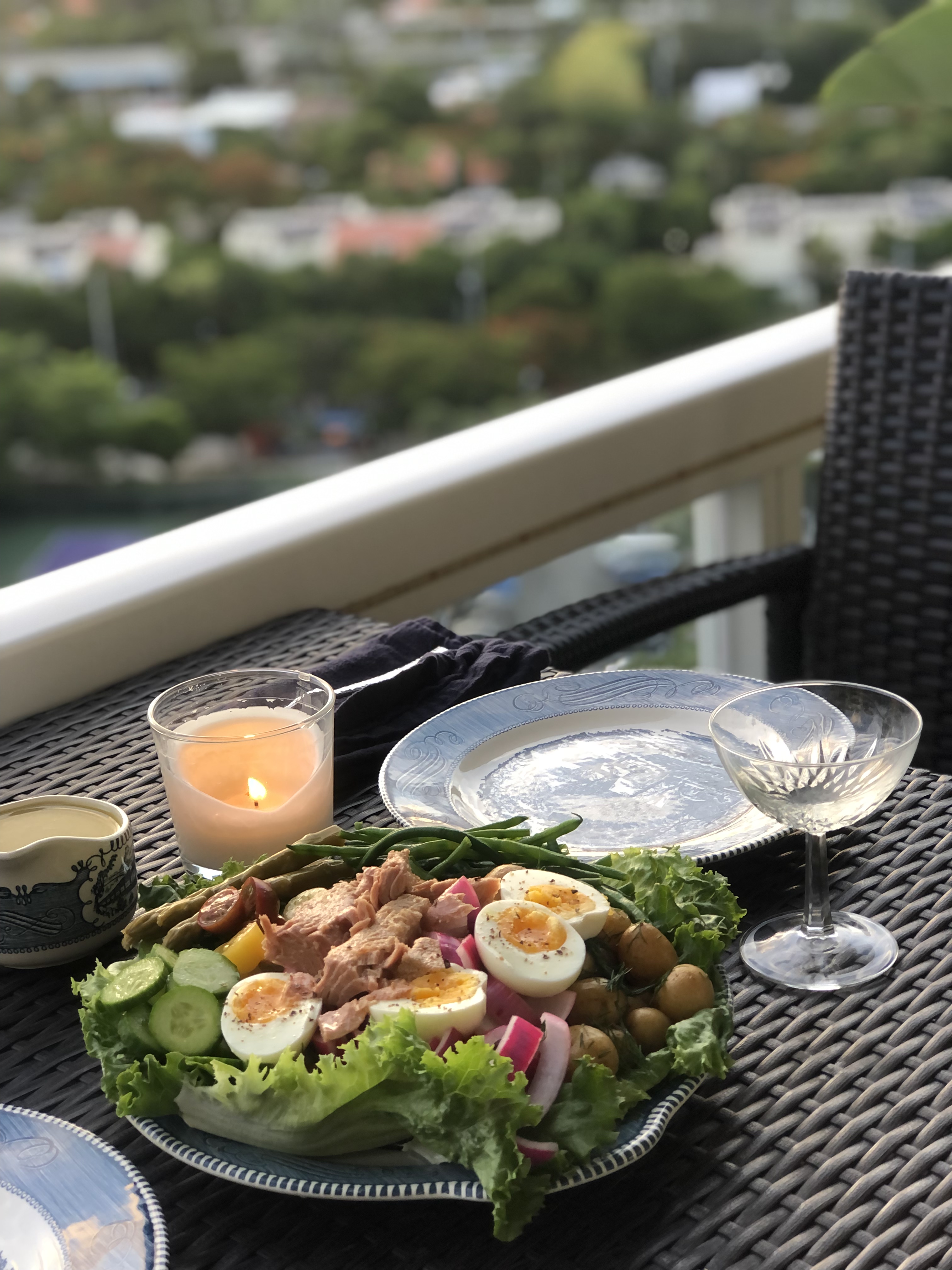 A tuna nicoise on a blue and white plate. The salad features green beans, pickled asparagus, medium boiled eggs, pickled red onions, baby potatoes with dill, sliced cucumber, sliced multicolor cherry tomatoes, green beans, romaine lettuce, and a pile of tuna. There is a candle lit on the table and a coupe glass of wine. A blurry view of Miami is visible in the background.