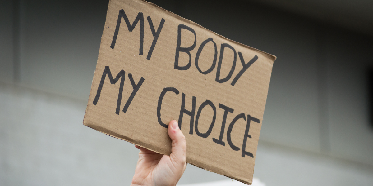 Against a white tiled background, a white person's hand holds up a cardboard sign that reads, 