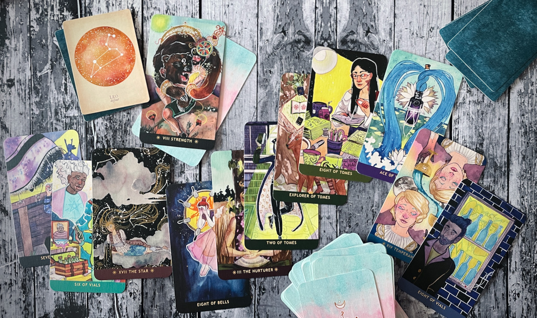cards from the numinous tarot are strewn together on a black wood table. They are of a variety of bright colors and there are aqua blue crystals at the edges.