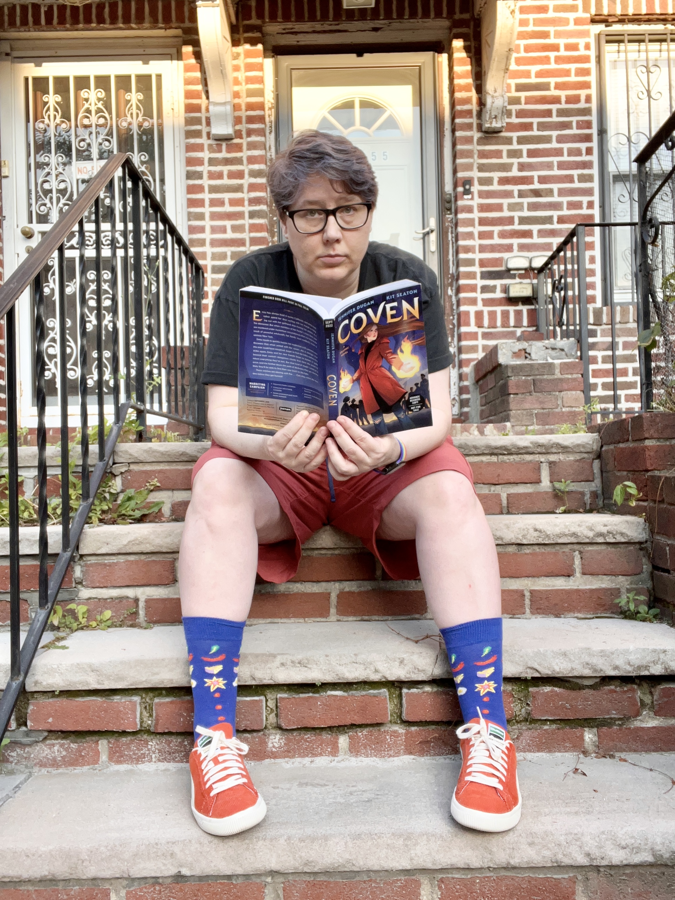 Heather is sitting on a stoop reading a book called Coven. She is a white woman with short gray hair, wearing a tee shirt, shorts, socks and sneakers.