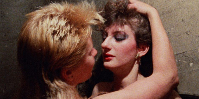 In a screenshot from Kamikaze Hearts, two women with mullets (one blonde, one brunette) are pressed up together against a wall. The blonde mullet has her arms grazing the shoulder and face of the brunette. It's the 1980s, with 1980s style bright make up included.
