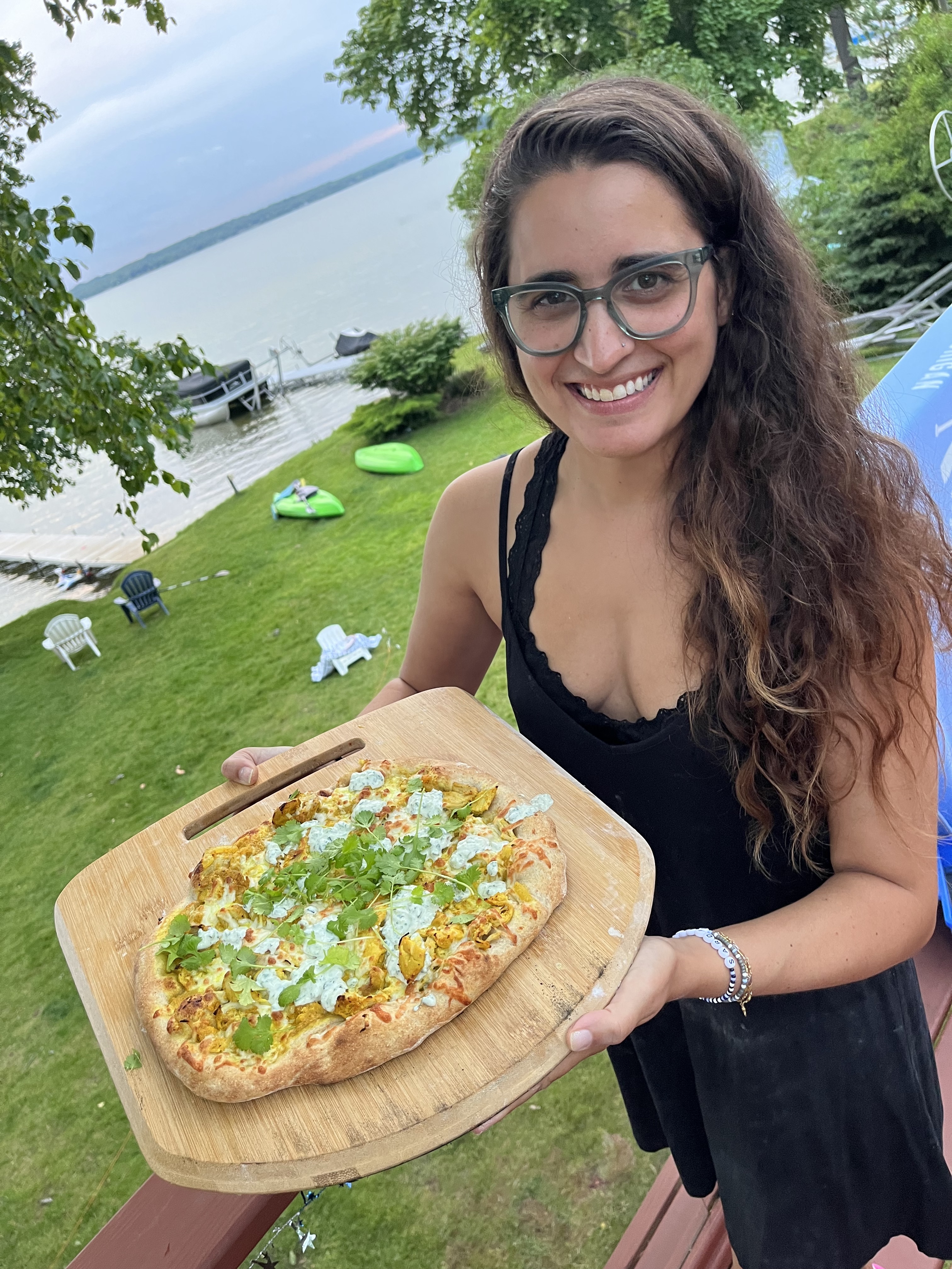 Kayla Kumari Upadhyaya holds a pizza topped with chicken makhni in front of a lake. She is wearing blue glasses and a black dress with a black lacy bra.