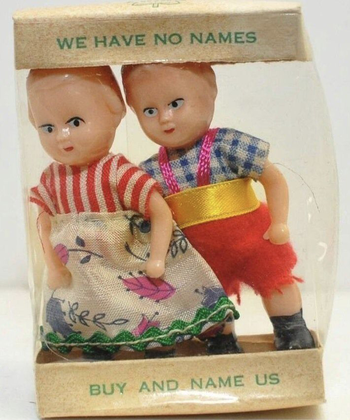 two creepy oldey timey white dolls in a plastic package with text that reads "we have no names. buy and name us" their eyes are somehow both vacant and alive